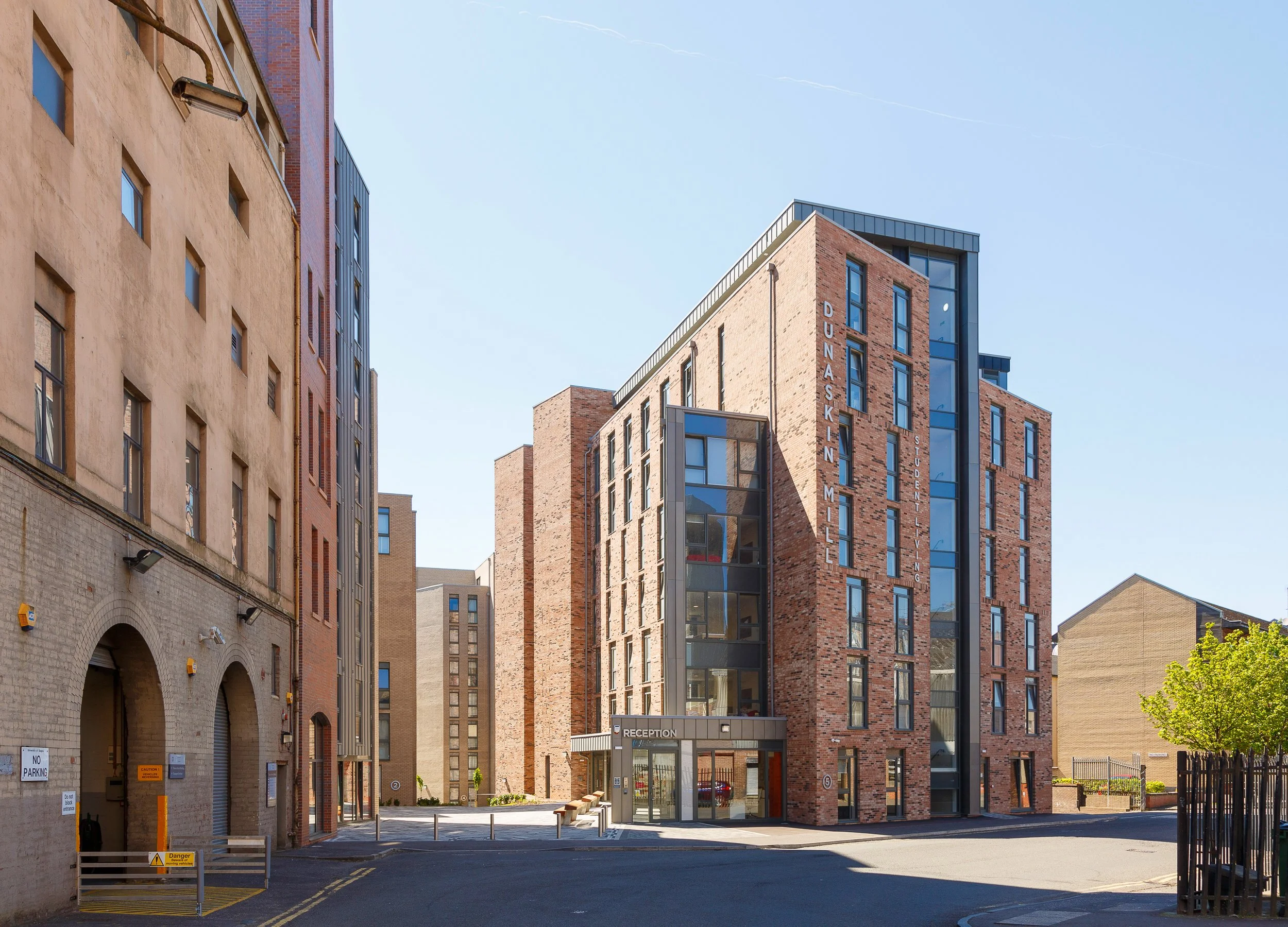 Modern multi-story brick building with glass accents and signage for Dunaskin Mill Student Living, located on a city street with surrounding buildings, trees, and a clear blue sky.