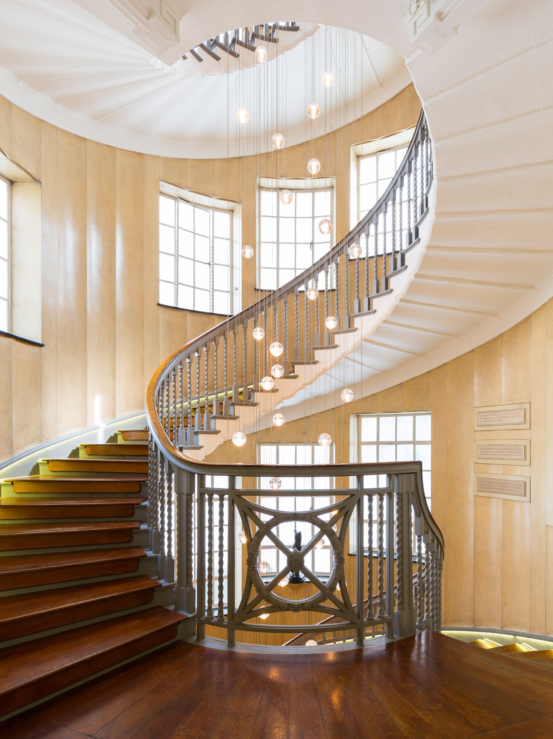 Interior view of a spiral staircase with wooden steps and decorative railing, illuminated by hanging spherical light fixtures, large multi-pane windows, and beige walls.