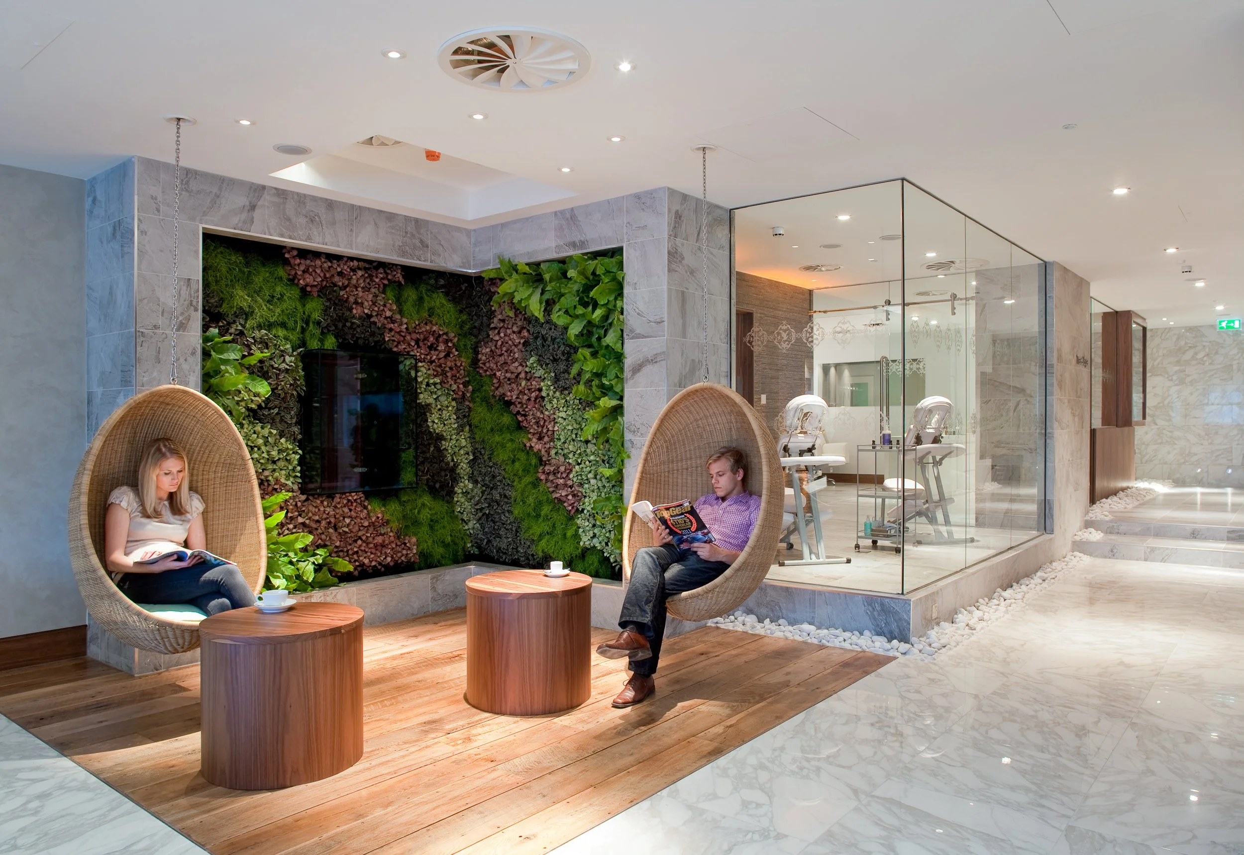 Interior of a spa or waiting area with two hanging woven chairs, a green wall, wooden tables, and a glass walled treatment room with massage chairs in the background.