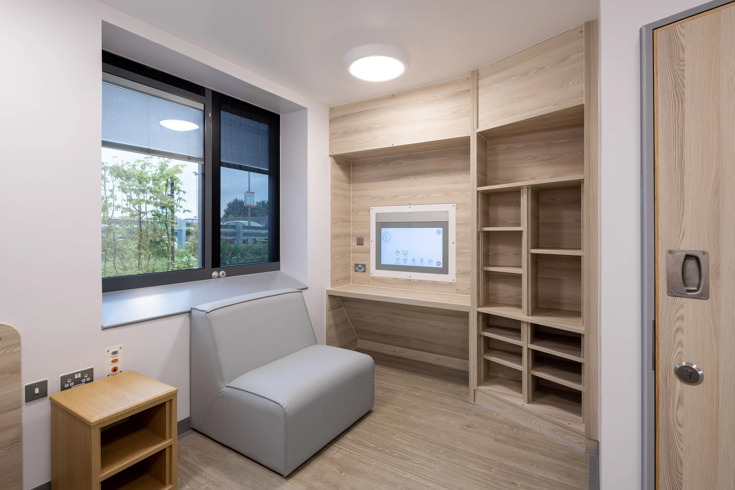 Empty room with wooden shelving, a window showing outside greenery, a small gray couch, and a built-in touchscreen on the wall.