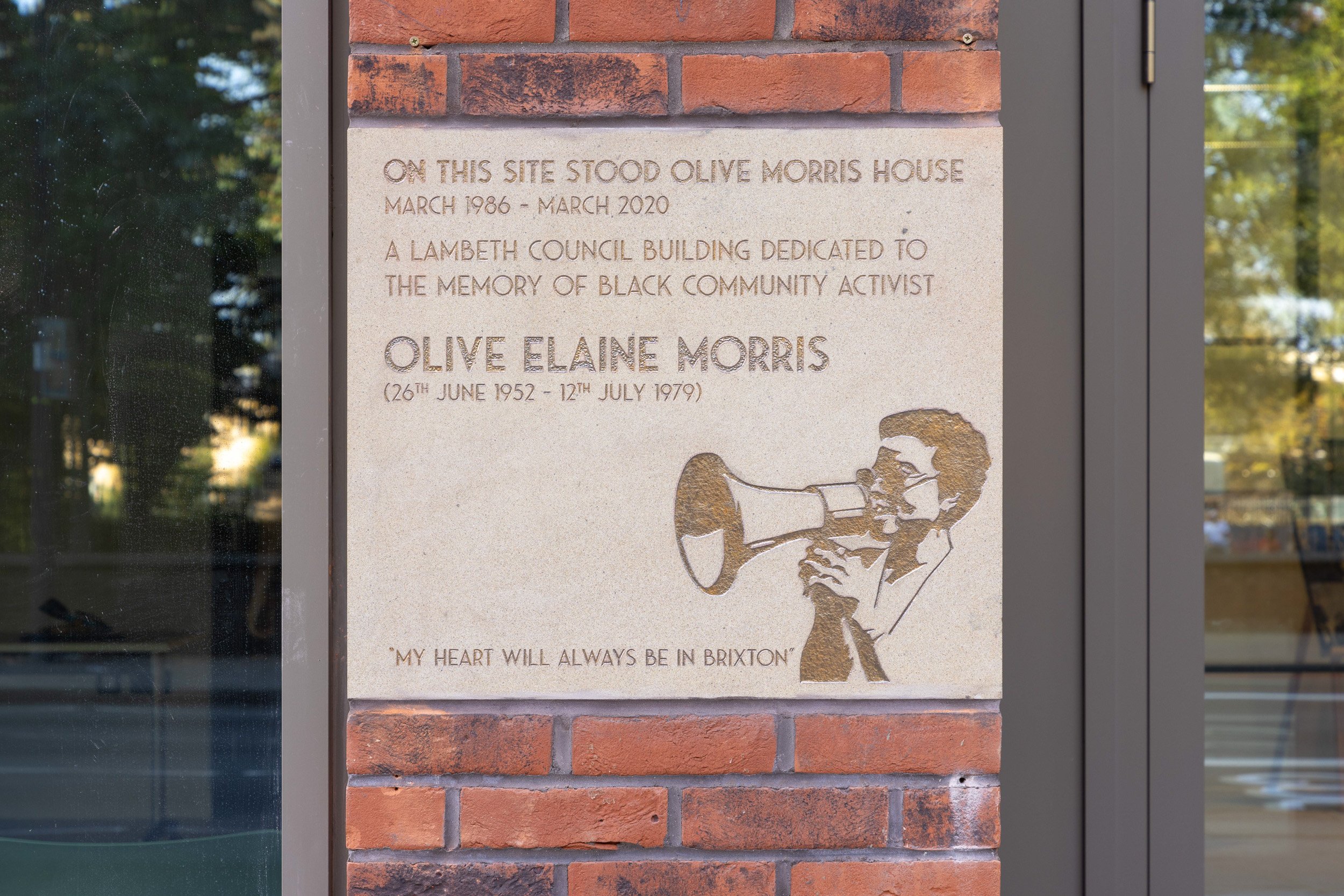 A memorial plaque dedicated to Olive Elaine Morris, a Black community activist, mounted on a brick wall. The plaque includes text about her life and a cutout illustration of her using a megaphone. The plaque text reads: "On this site stood Olive Morr