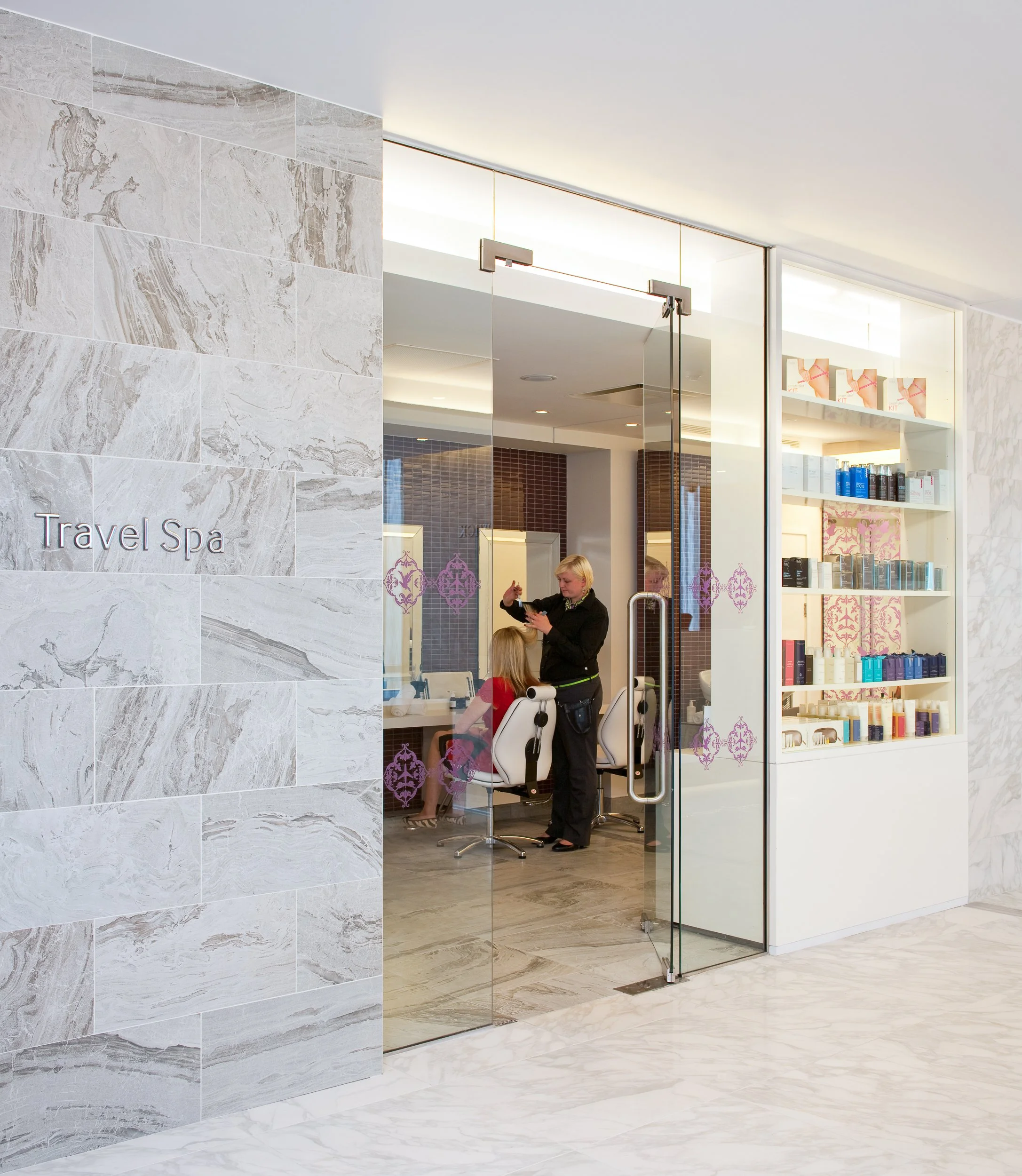 Exterior view of a Travel Spa salon with a glass door and pink decorative designs, with a stylist working on a client inside.