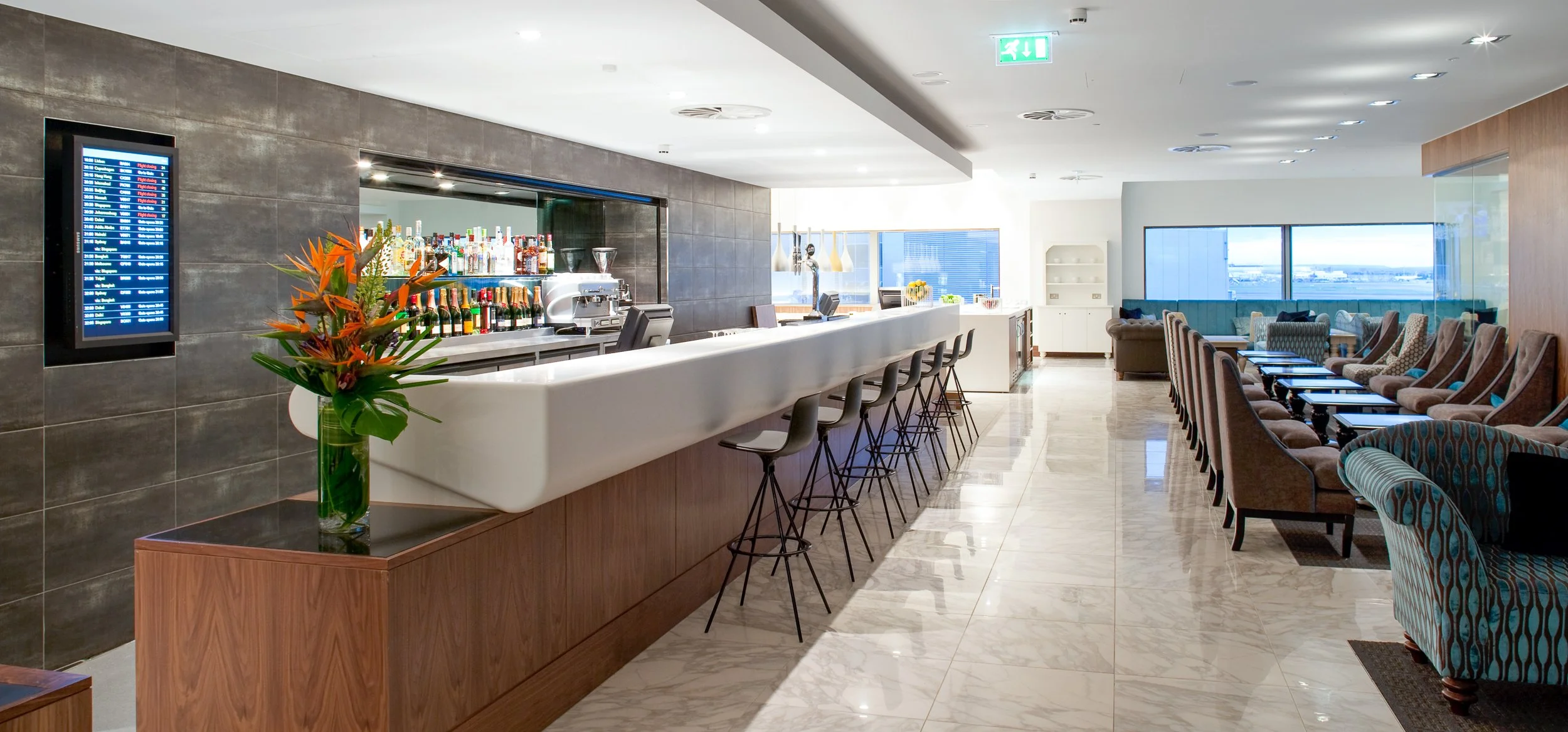 Modern hotel bar and lounge area with a white curved bar counter, black bar stools, a colorful flower arrangement on a small table, and a seating area with patterned armchairs and sofas near large windows with city and river views.