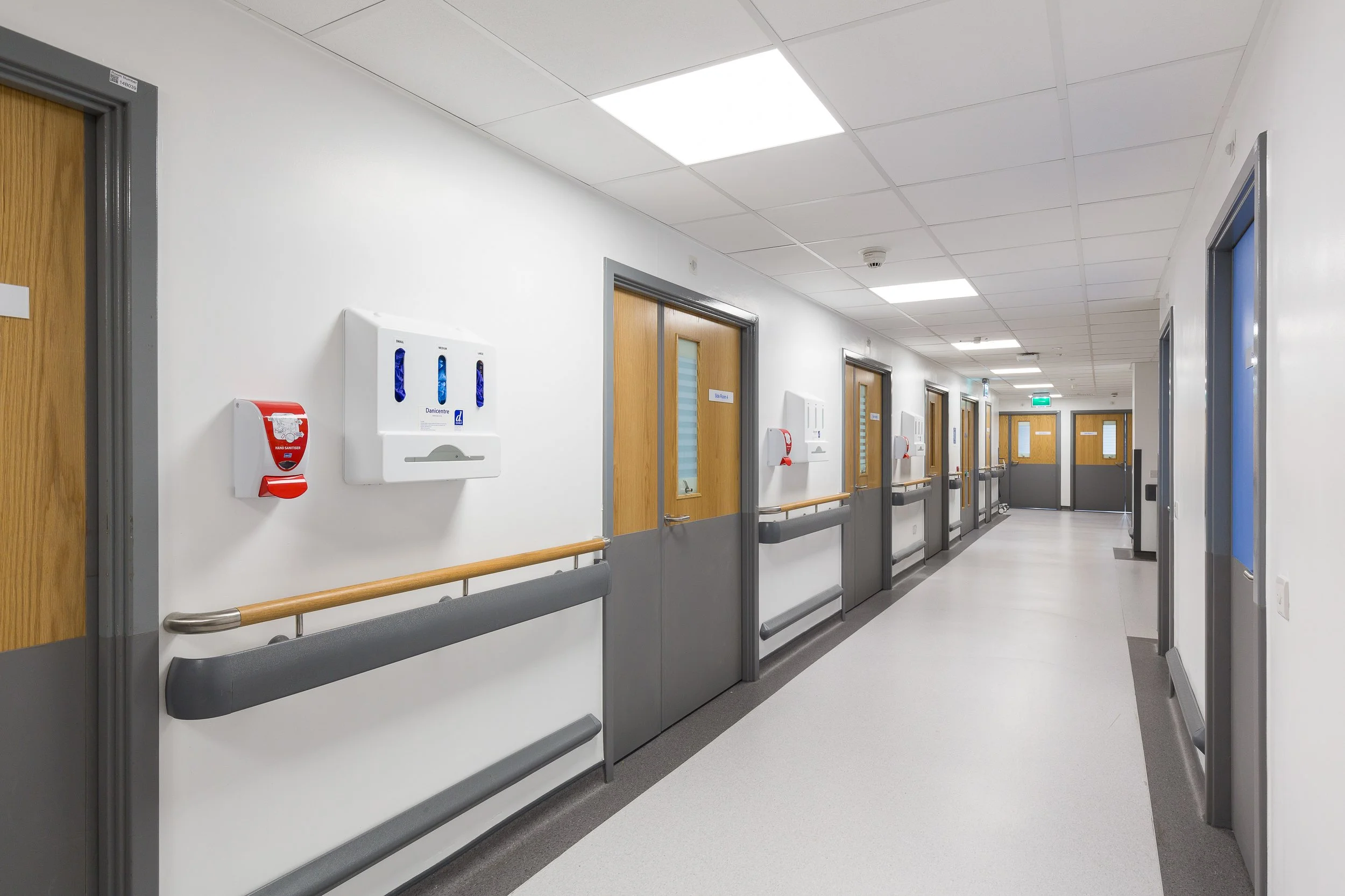 Empty hospital corridor with multiple patient rooms, wooden doors with windows, white walls, ceiling lights, fire alarm, hand sanitizer dispenser, and grab bars along the wall.