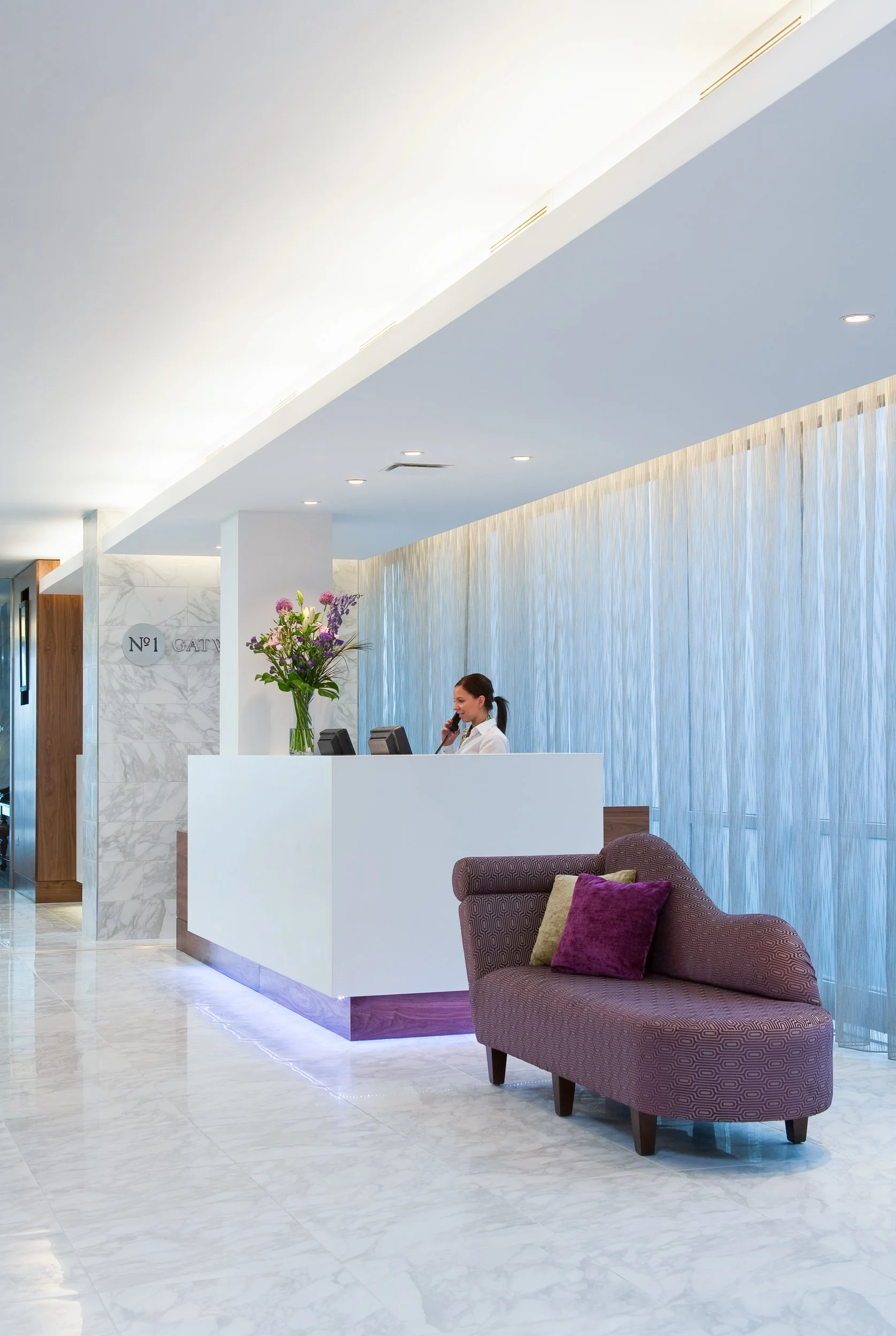 Hotel reception area with a white counter, a woman speaking on the phone, a vase of purple flowers, and a purple upholstered sofa with pillows.