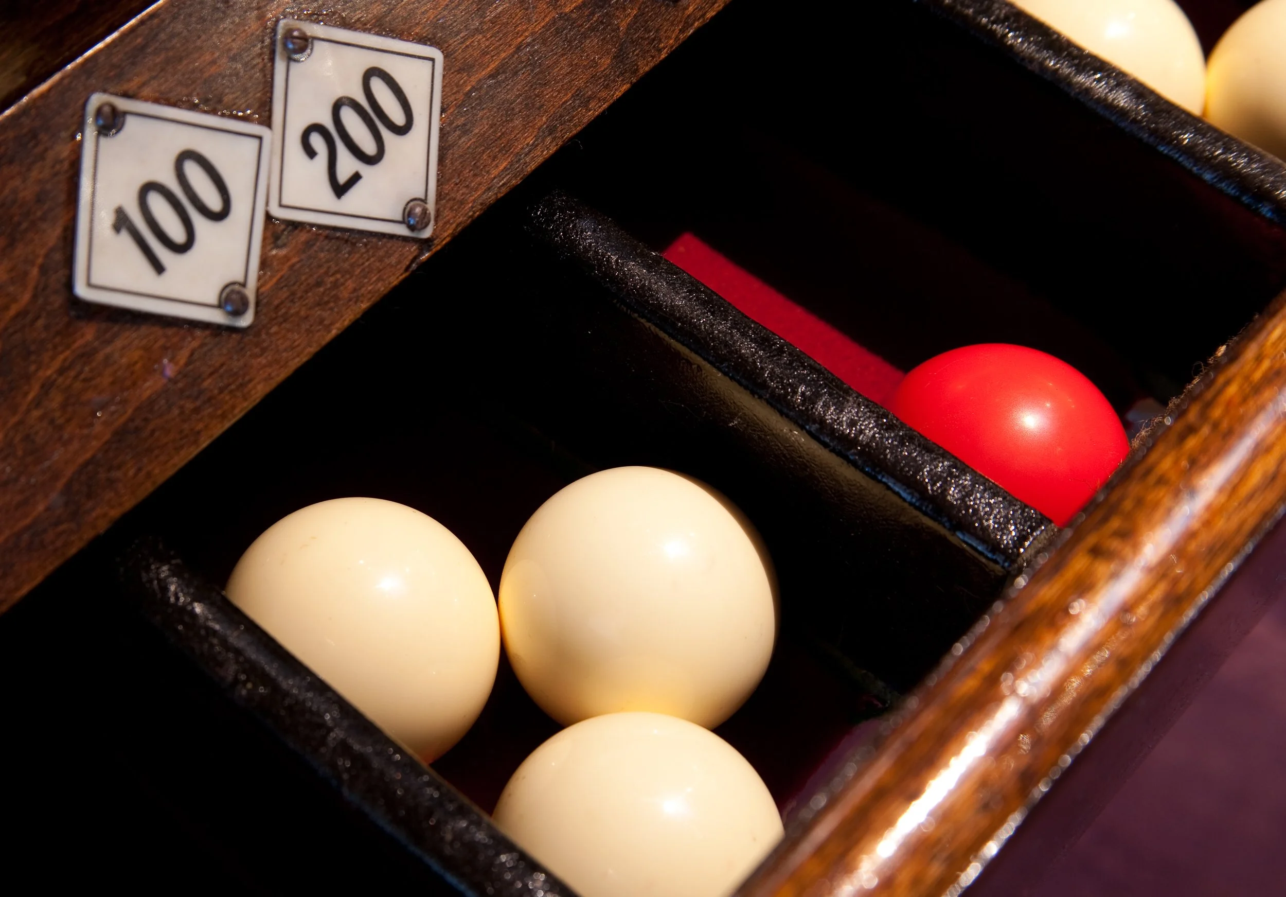 A billiard ball rack holding white balls with one red ball, inside a wooden box with betting chips labeled 100 and 200.