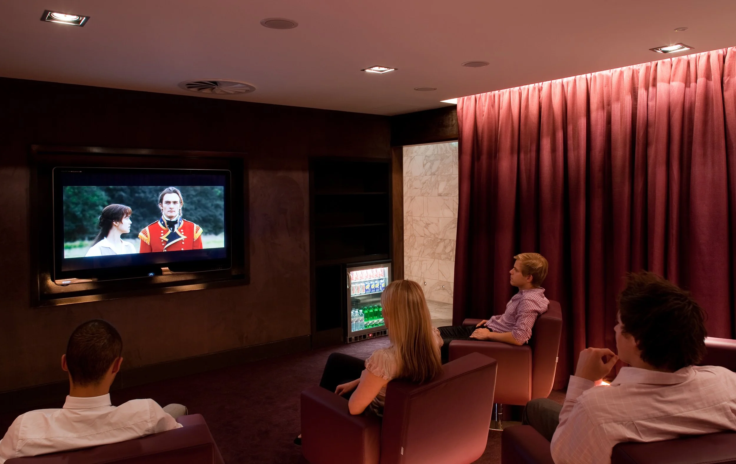 Four people sitting in a dark room watching a TV displaying a scene with a man and woman outdoors, with maroon curtains to the right and a small fridge underneath the TV.