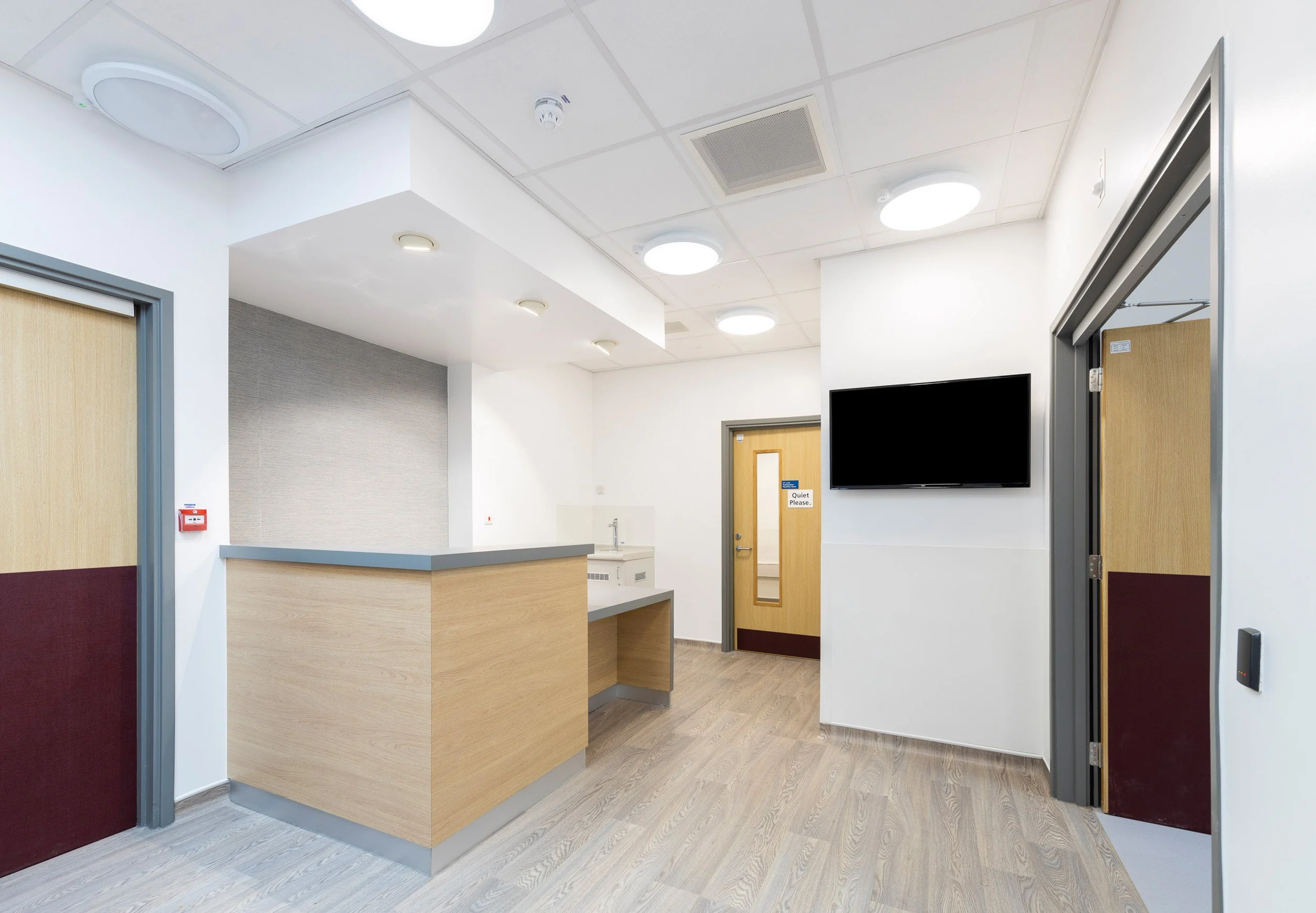 A hospital reception area with a wooden reception desk, a wall-mounted TV, a door marked 'Quiet Please', and an open doorway leading to another room. The room has ceiling lights, a smoke detector, and a hand sanitizer sink.