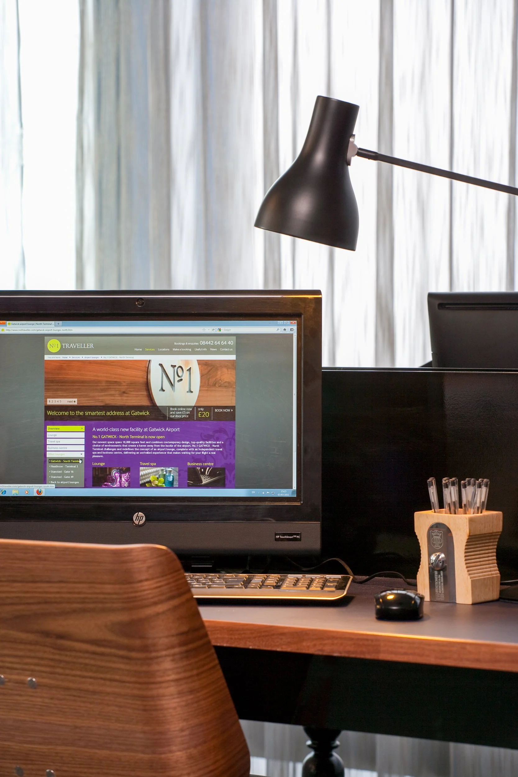 Computer monitor on a wooden desk displaying a travel website, with a black desk lamp overhead and a wooden pen holder with pens on the right.