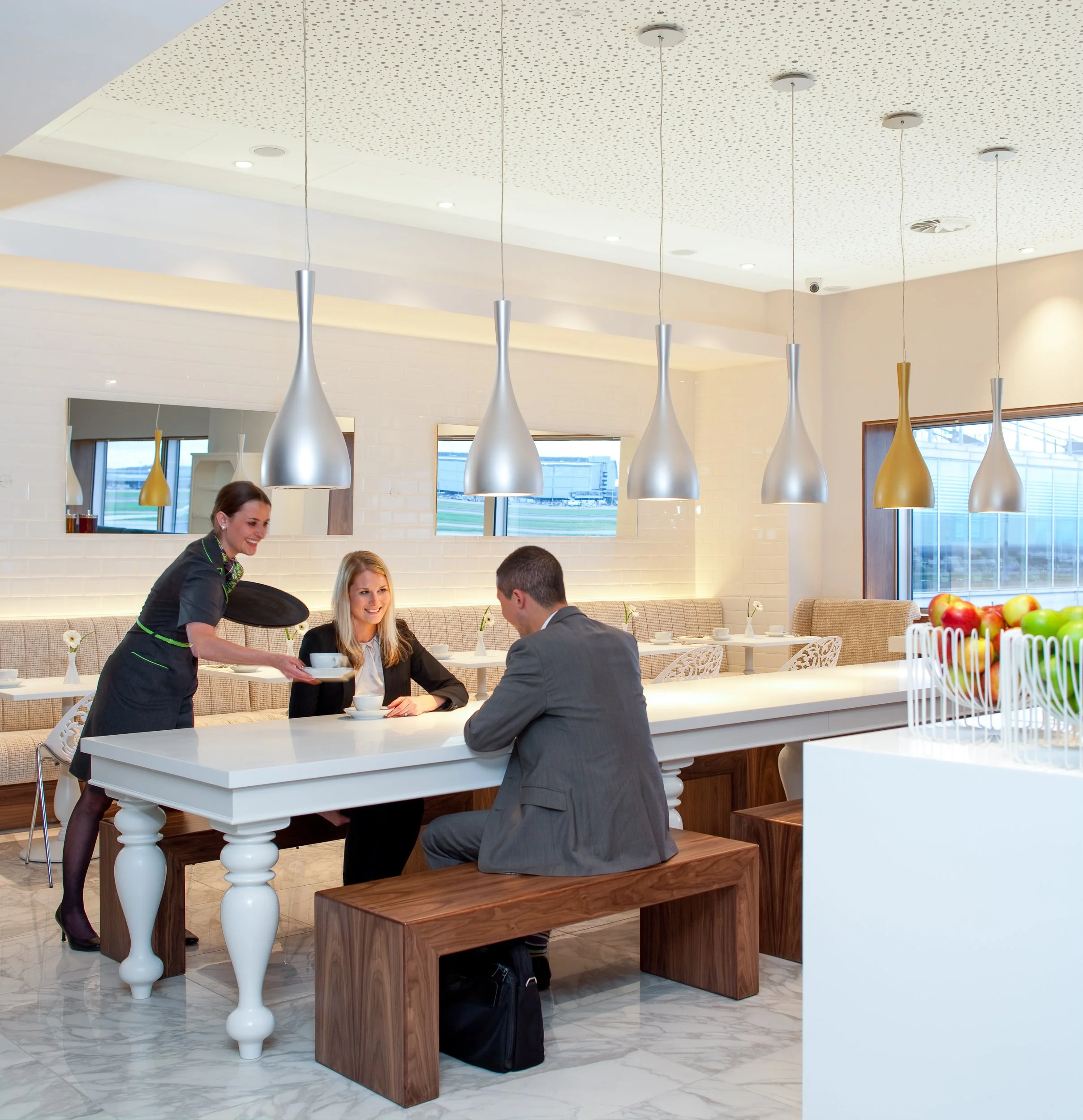 A waiter serving tea to a woman sitting at a table in a modern cafe or restaurant with pendant lights and large windows.