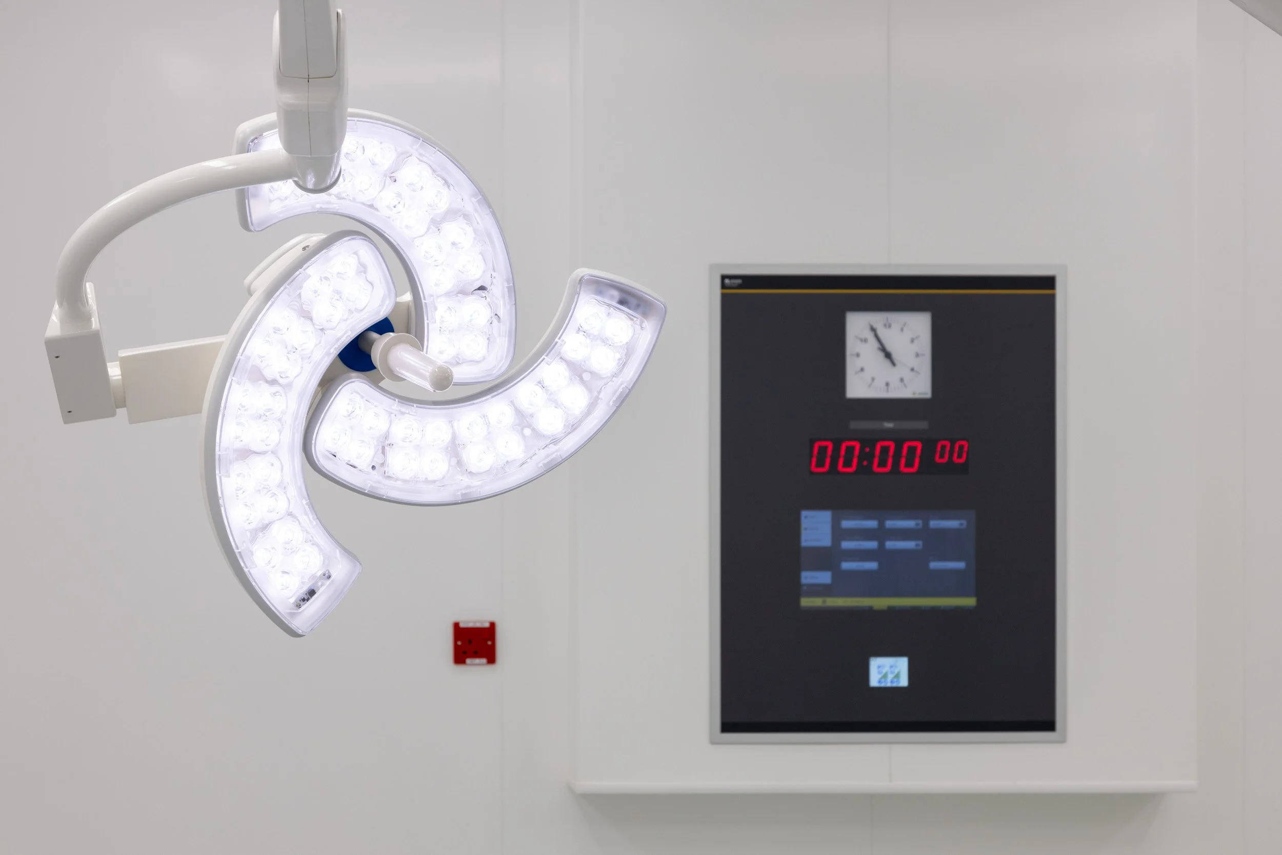 An overhead surgical light in a sterile operating room with a digital timer and clock on the wall.
