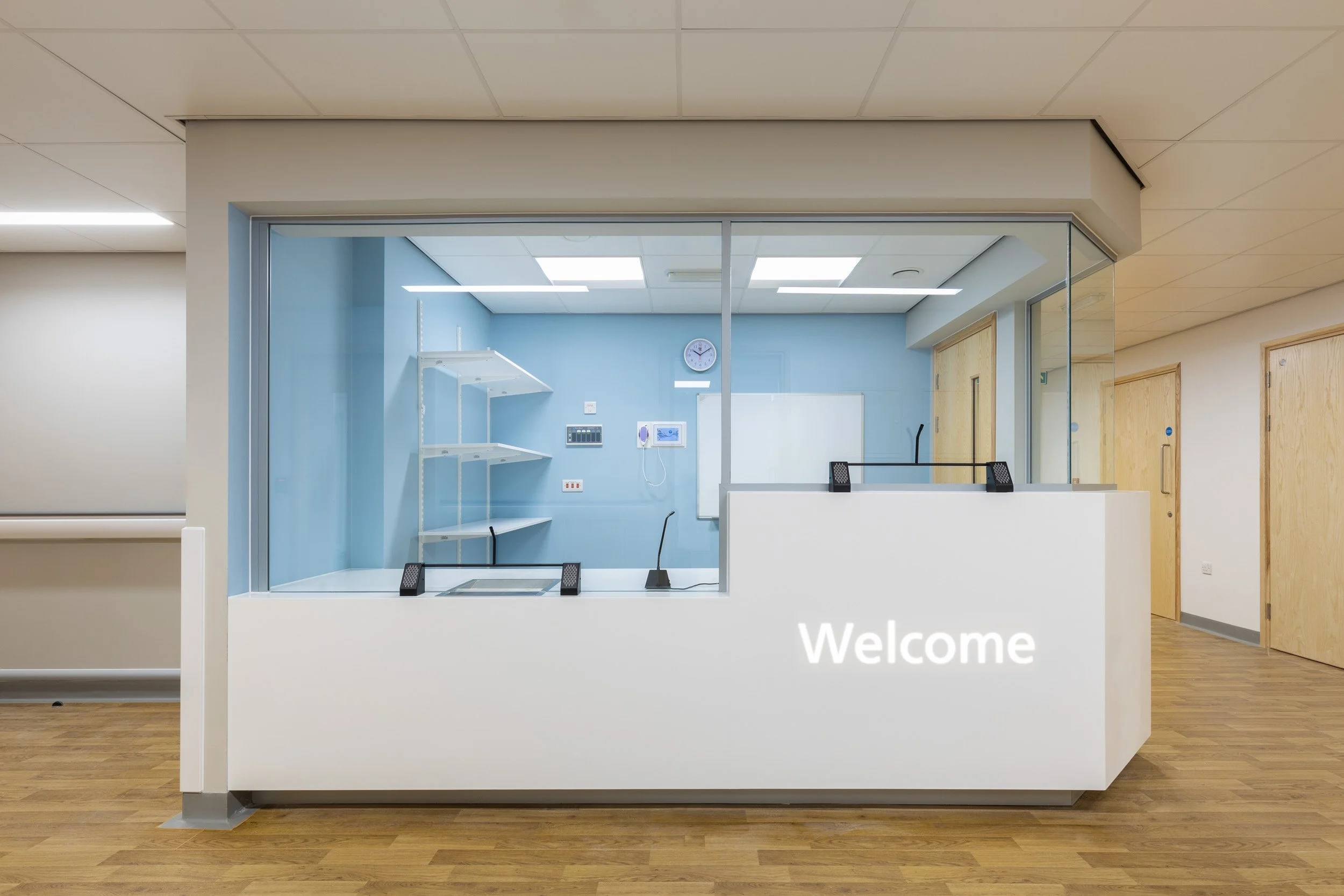 Hospital reception desk with a white counter and the word "Welcome" written on it. Behind the glass partition, there is a blue wall, a whiteboard, monitor, clock, shelves, and some equipment. The floor is wooden, and there are several doors on the ri
