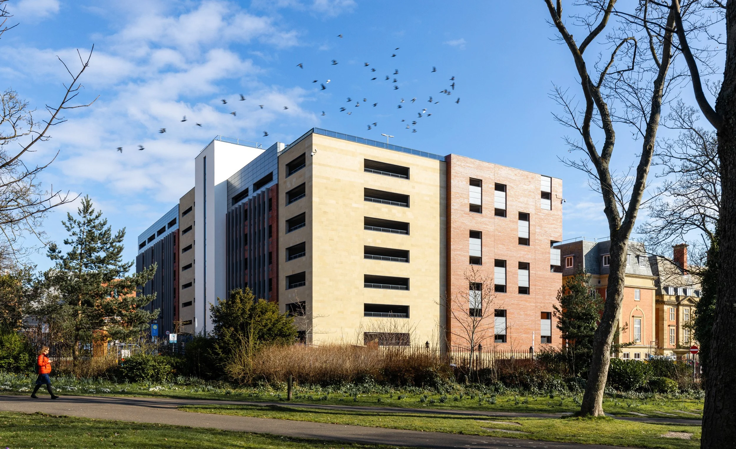 A modern multi-story apartment building with a mix of beige, red brick, and white exteriors, set against a blue sky with bird flight, trees, and a person walking on a path in a park.