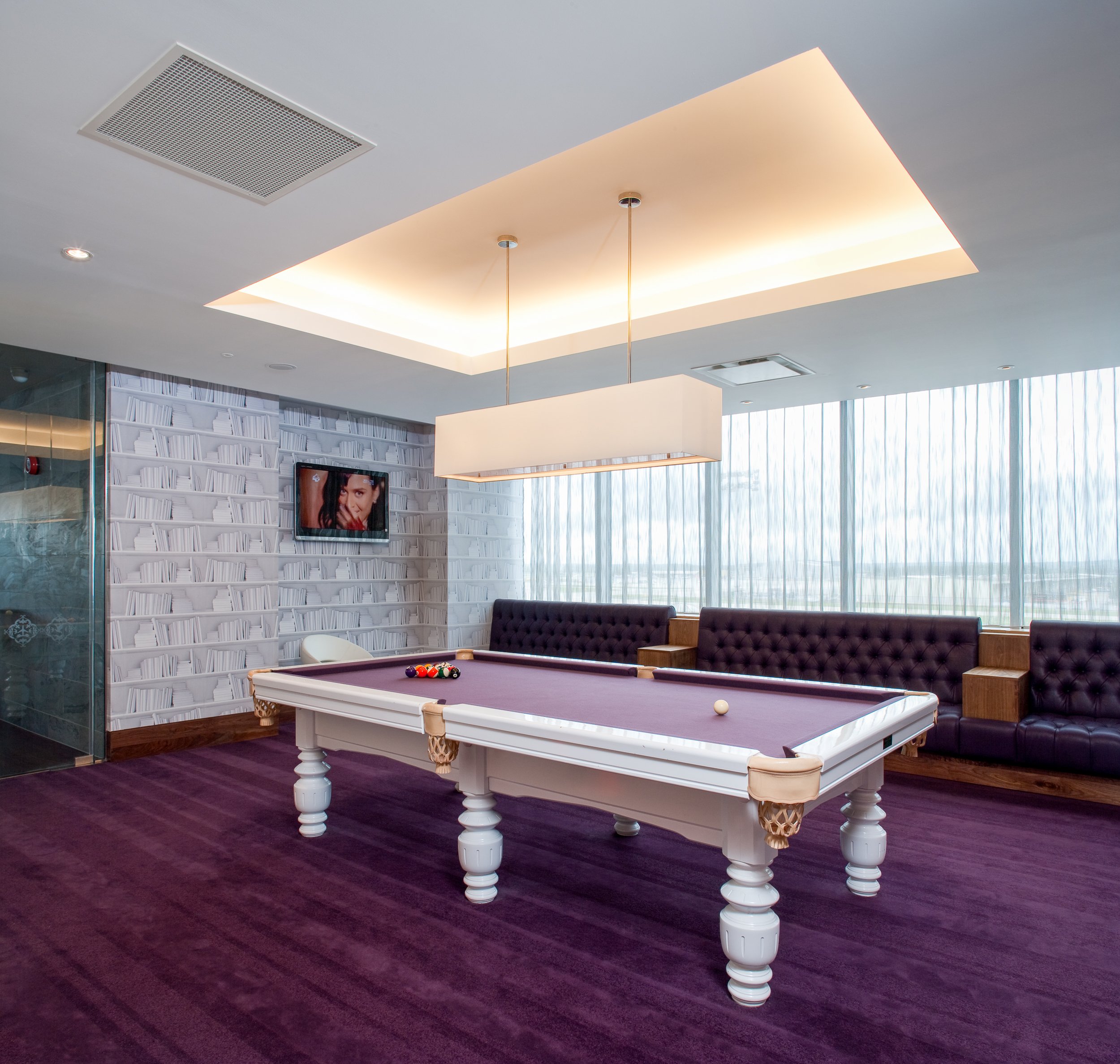 Indoor lounge with a purple-felt pool table, surrounded by purple cushioned seating and large windows with blinds, featuring a wall-mounted television and modern ceiling lighting.