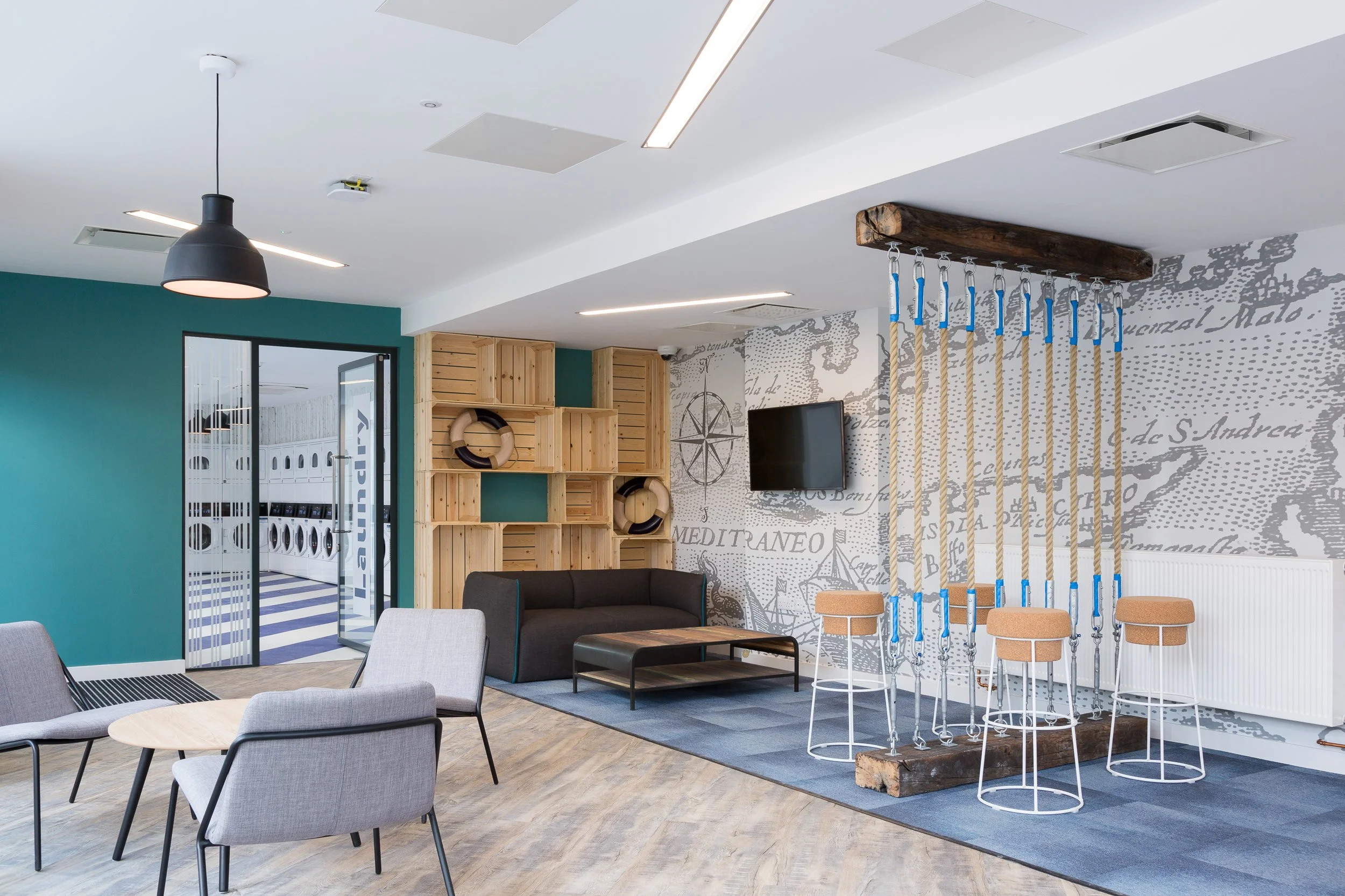 Indoor lounge area with chairs, a sofa, a wall-mounted TV, and a wooden shelving unit. A wall with a map design and an indoor climbing or swinging apparatus made of wooden beams and ropes. Laundry room visible through an open door in the background.