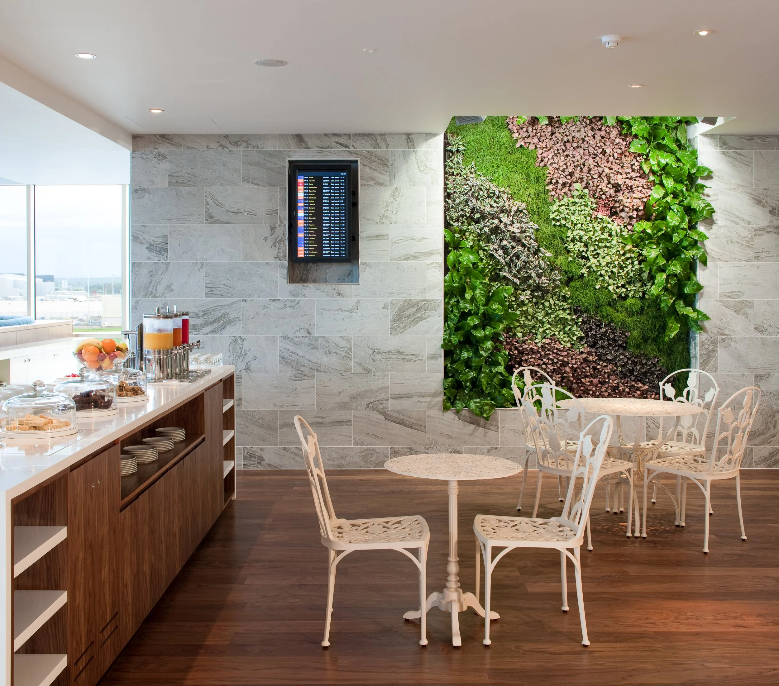 A hotel breakfast area with a buffet table on the left, a digital flight information display on the wall, and a green wall art installation on the right, with white tables and chairs in the foreground.