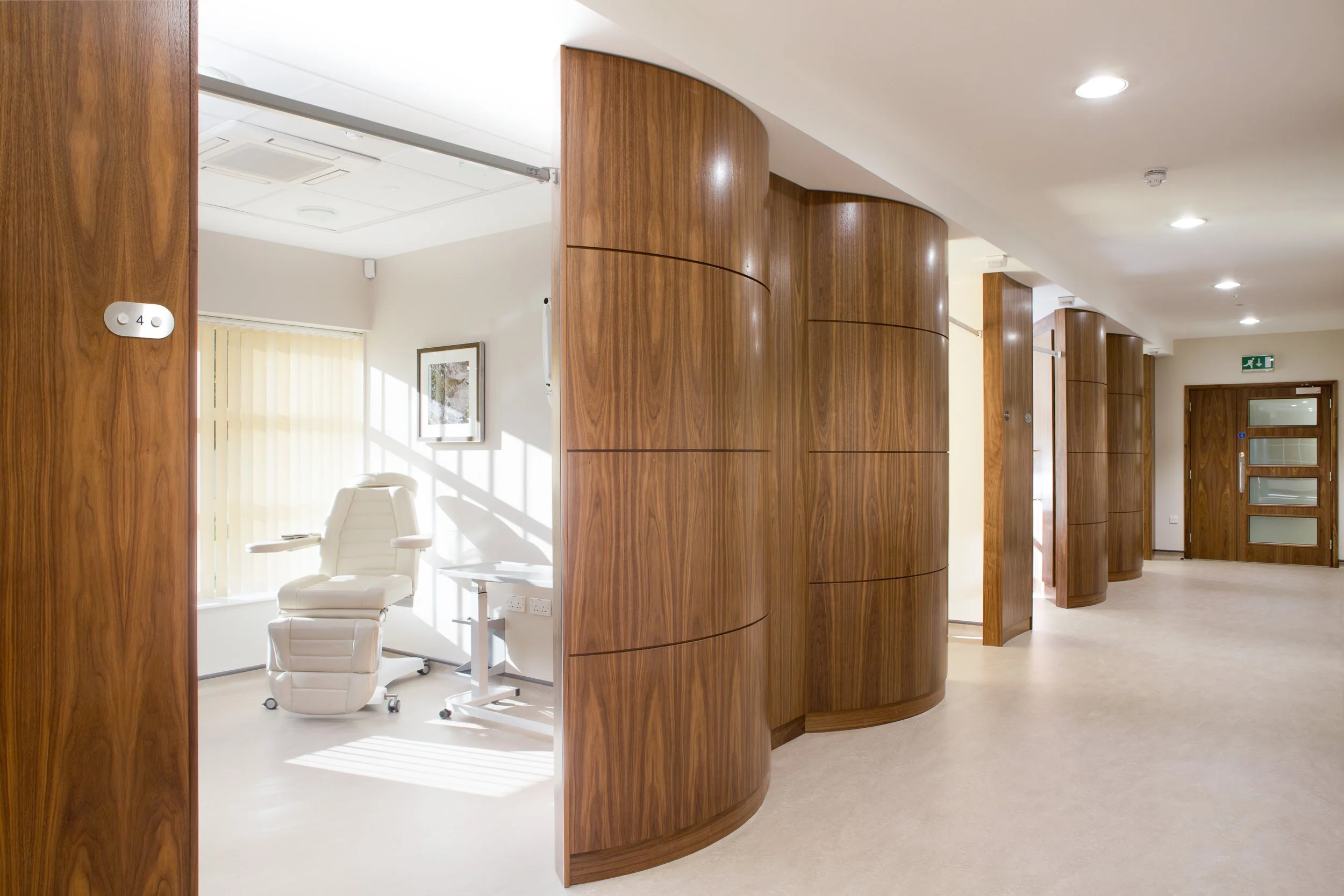 Empty hospital or clinic room with a white reclining chair, a small table, and a framed picture on the wall. The room is bright with natural light, and the hallway features curved wooden panels, a closed wooden door, and ceiling lights.