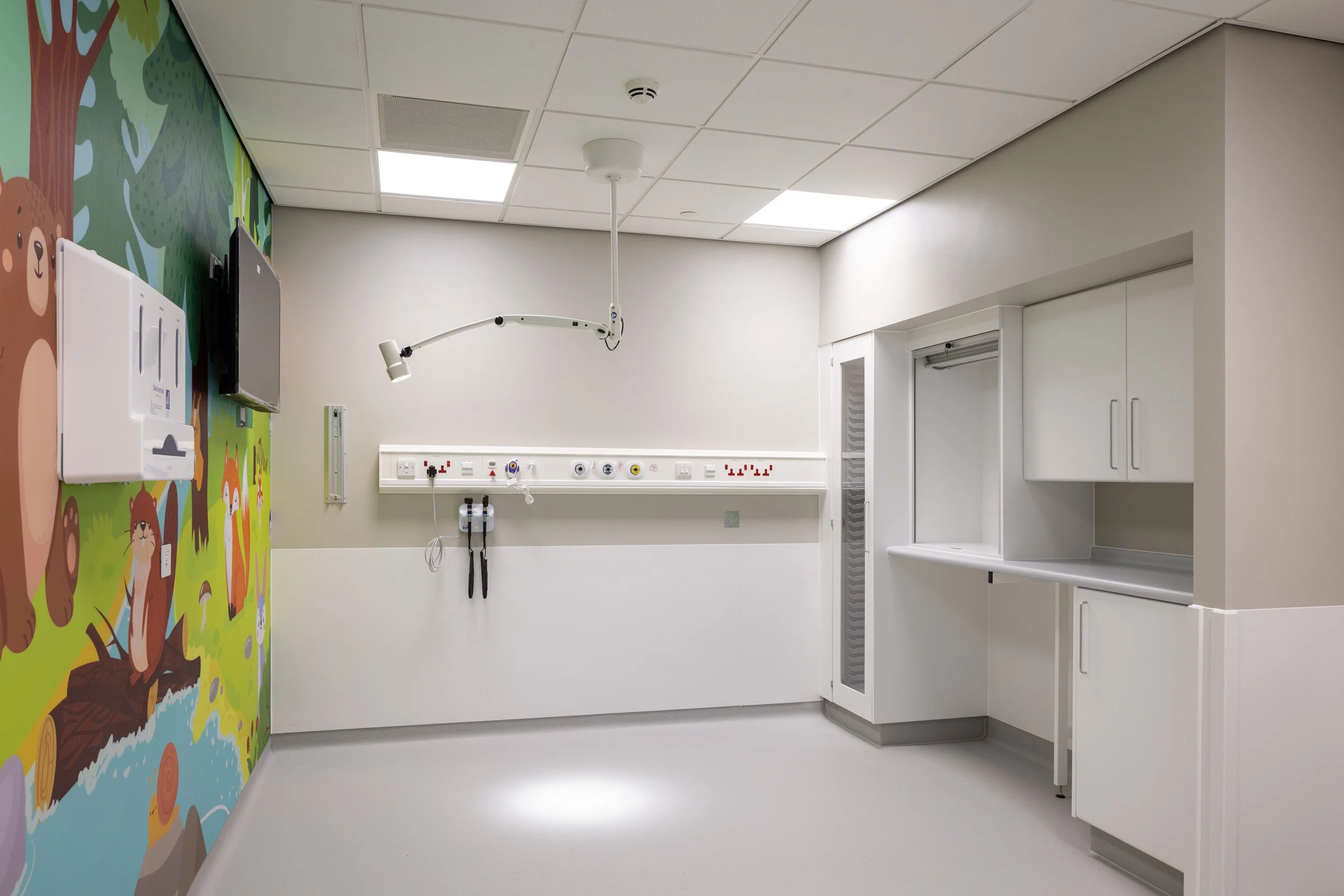 Empty hospital pediatric examination room with colorful bear-themed wall mural, medical equipment, and cabinets.