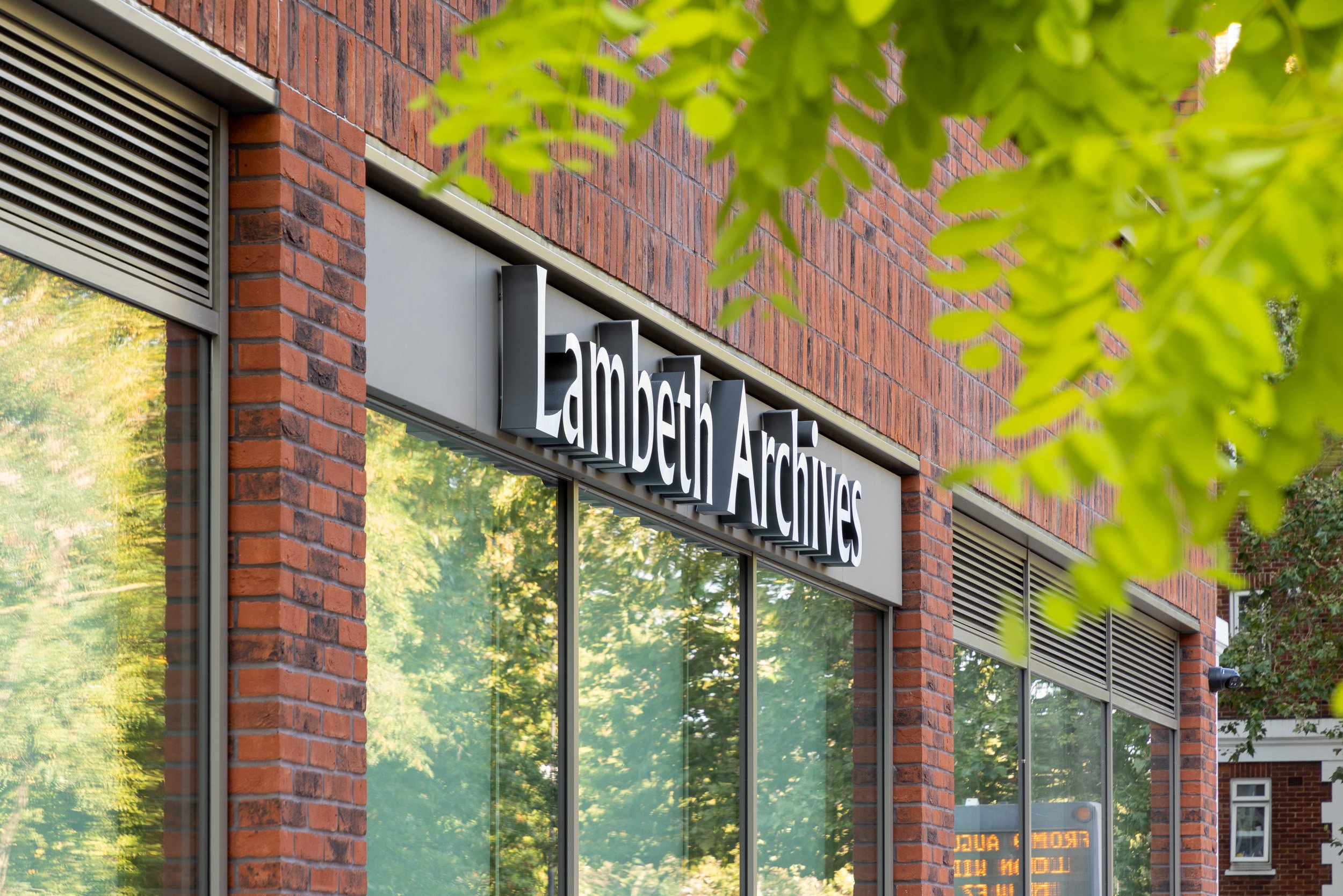 The exterior of Lambert Archives building with large windows and a sign on the brick wall, surrounded by green tree leaves.