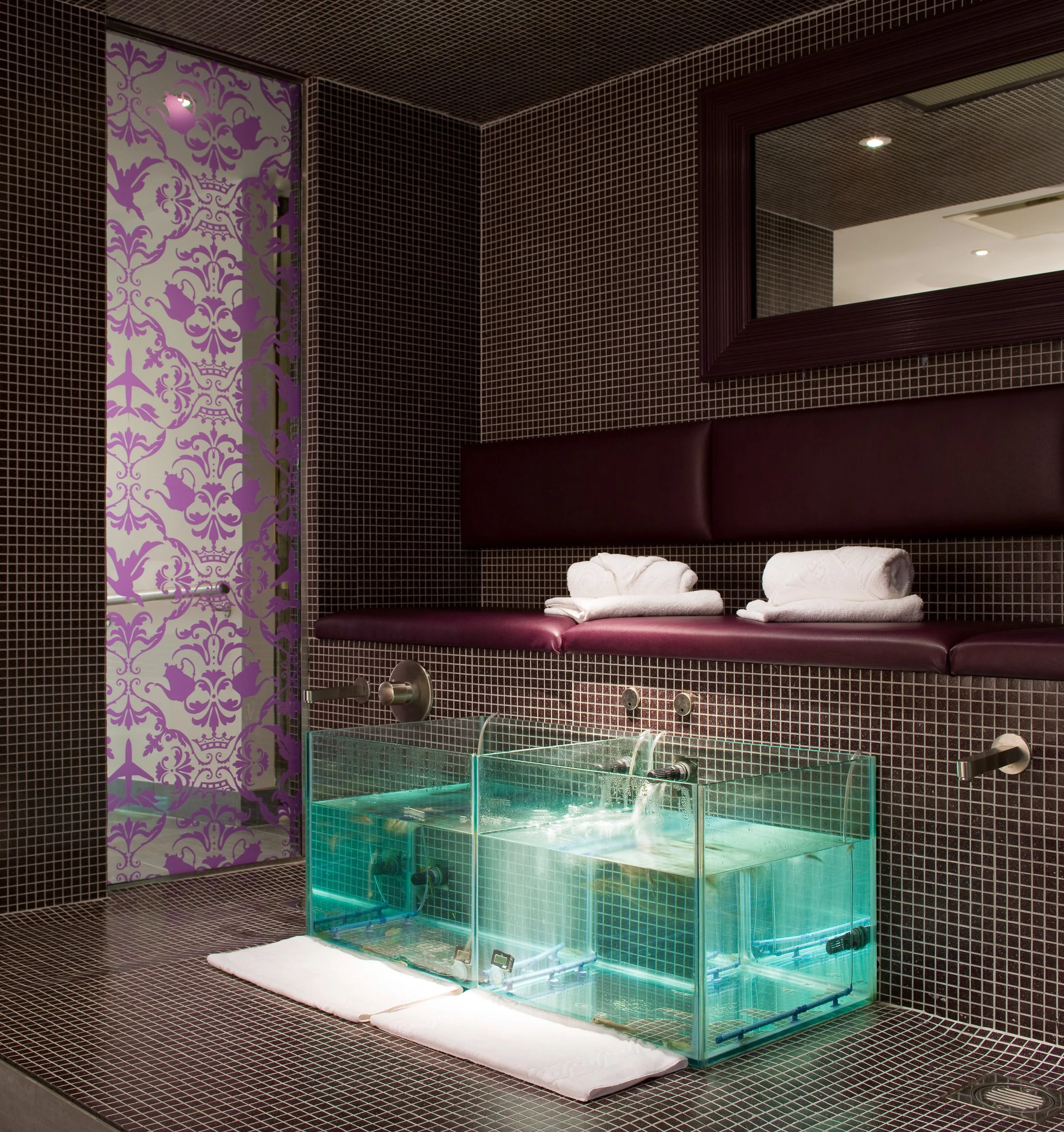 Luxury spa room with a glass-walled foot bath, folded white towels, purple padded bench, decorative purple pattern on the door, dark mosaic tiled walls and floor, and a large mirror.