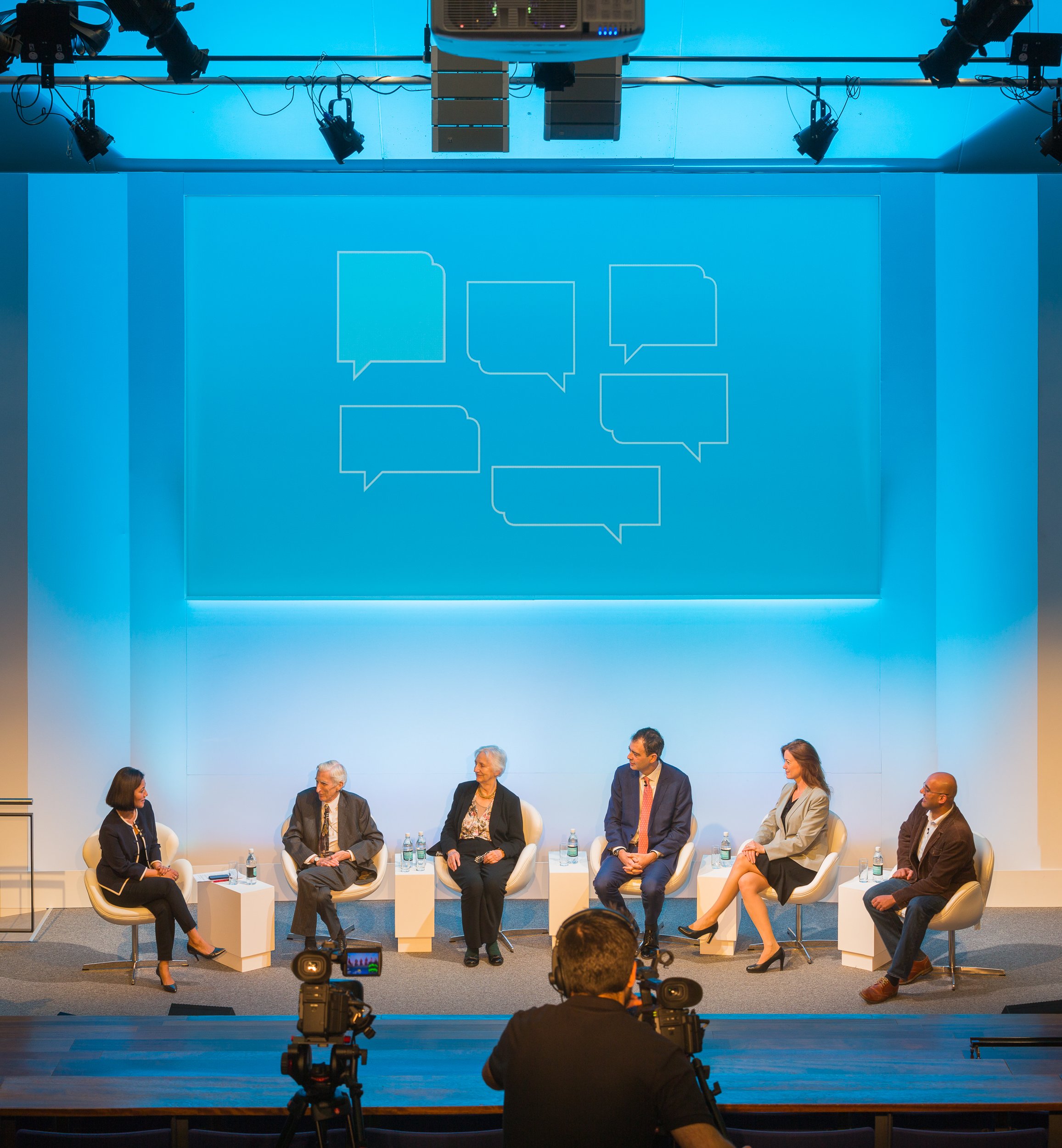Six people seated on stage in a panel discussion, with a projection screen behind them displaying speech bubbles and a camera operator filming the event.