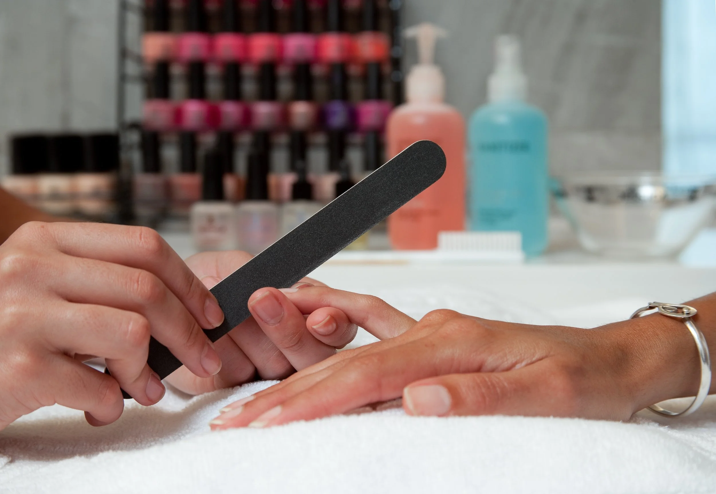 Manicurist filing a client's fingernail during a nail care session, with nail polish bottles and manicure tools in the background.