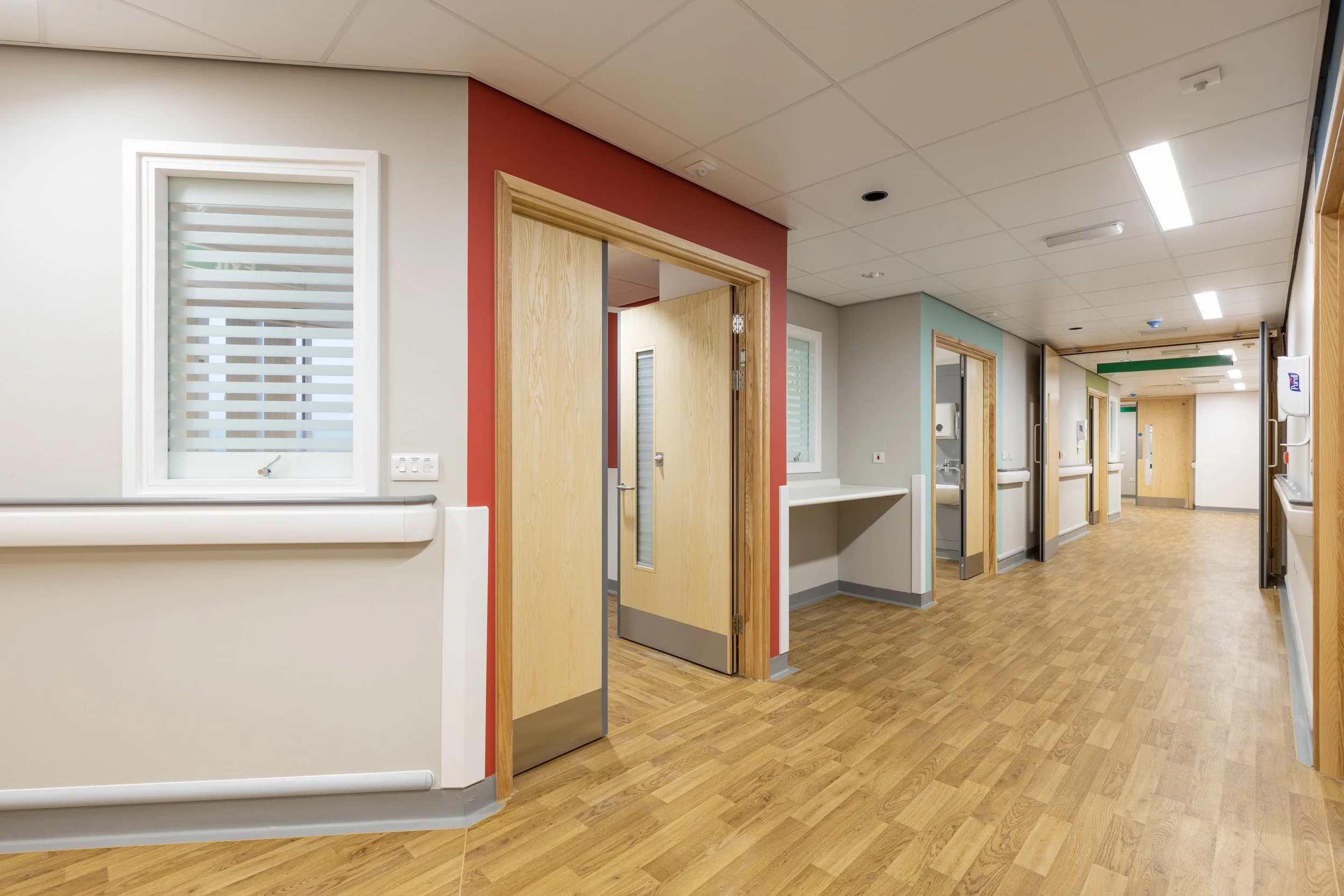 Empty hospital corridor with wooden floors, multiple doorways, handrails, and medical equipment.