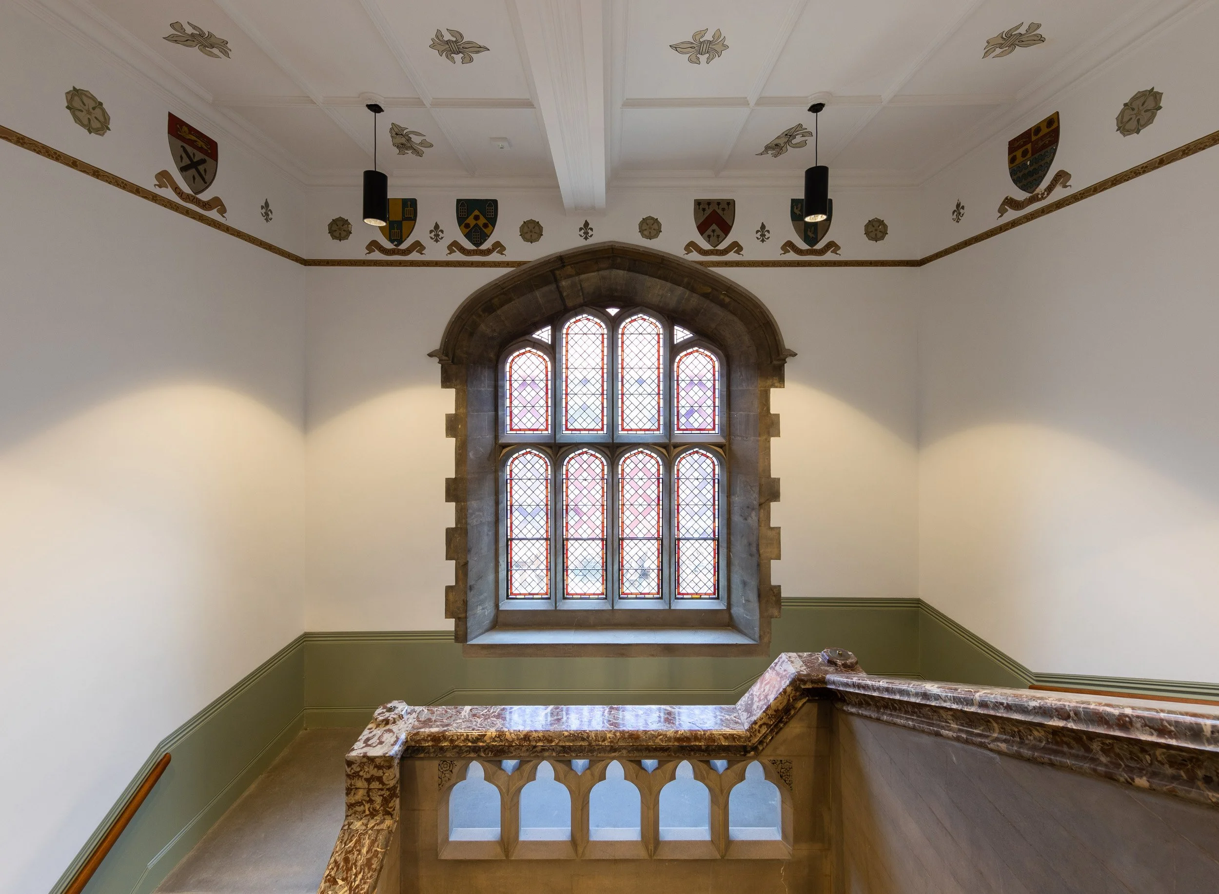 Interior of a historic building with a large arched stained glass window, featuring a stone frame and decorative glass patterns, with a marble railing in the foreground and coats of arms painted on the upper walls.