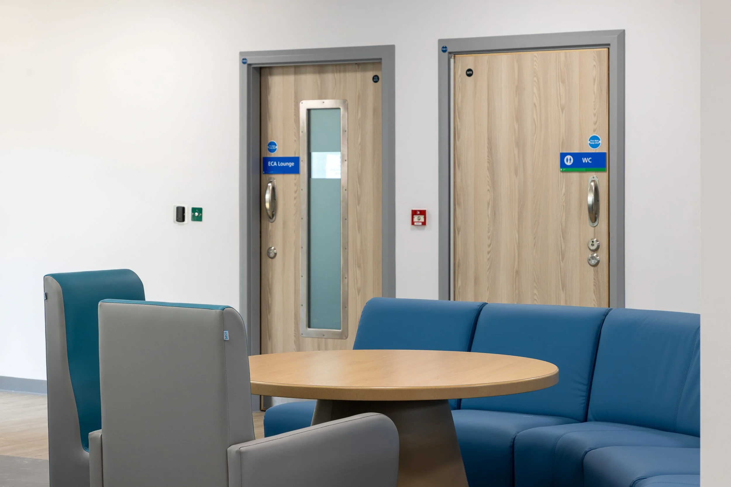 Waiting area with a round wooden table, blue and gray chairs, and two wooden doors labeled 'ECA Lounge' and 'WC' in an indoor setting.