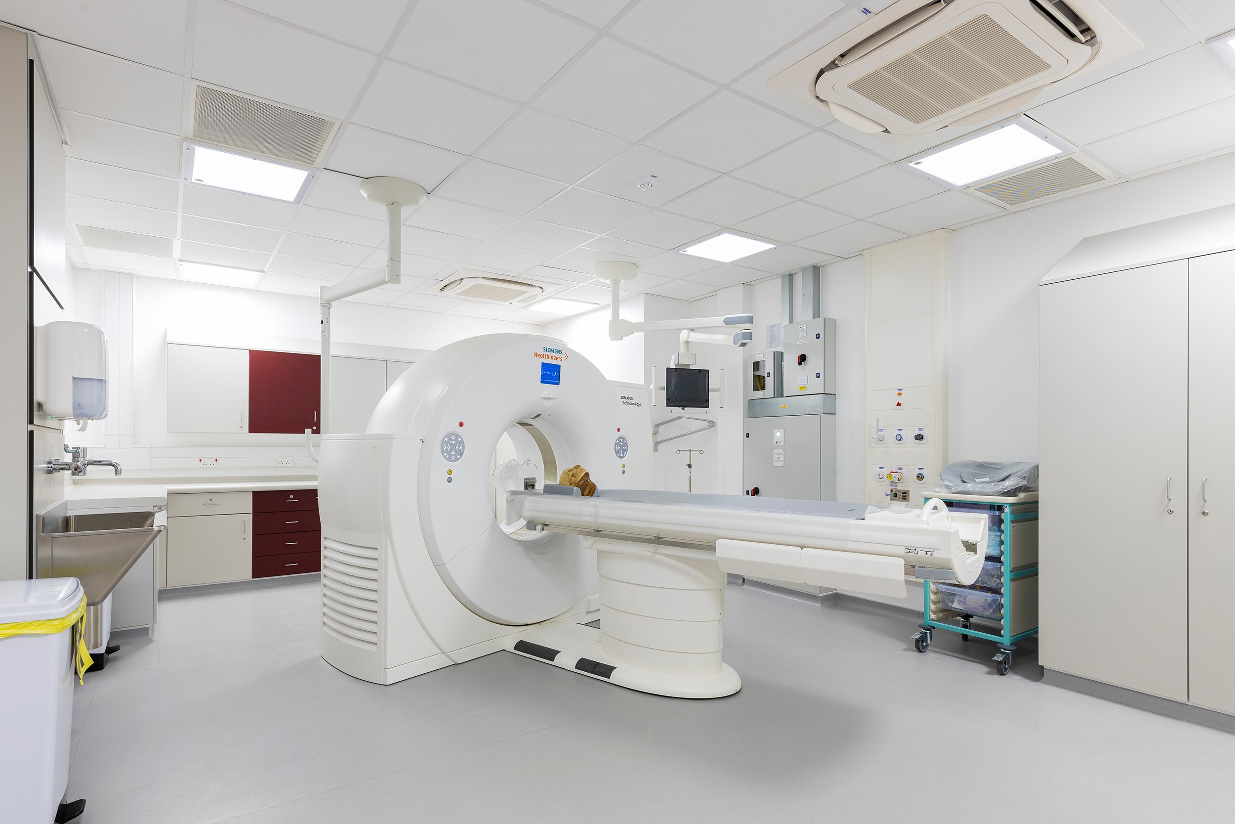 Empty hospital room with a large medical imaging machine, likely a CT scanner, in the center. The room has white walls, ceiling tiles, and various medical cabinets and equipment.