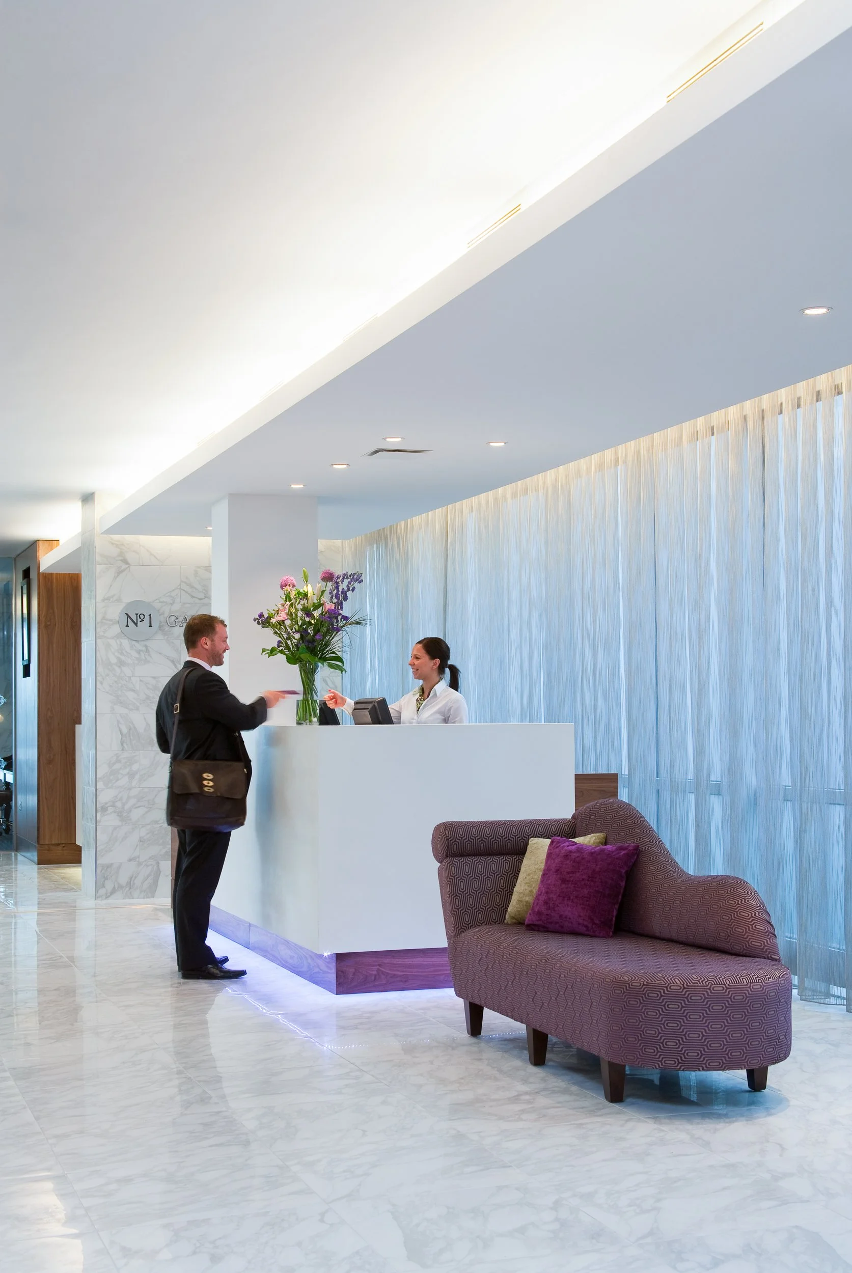 Hotel reception desk with a man checking in and a receptionist smiling behind the counter, decorated with a large vase of flowers, with a purple sofa and cushions nearby.
