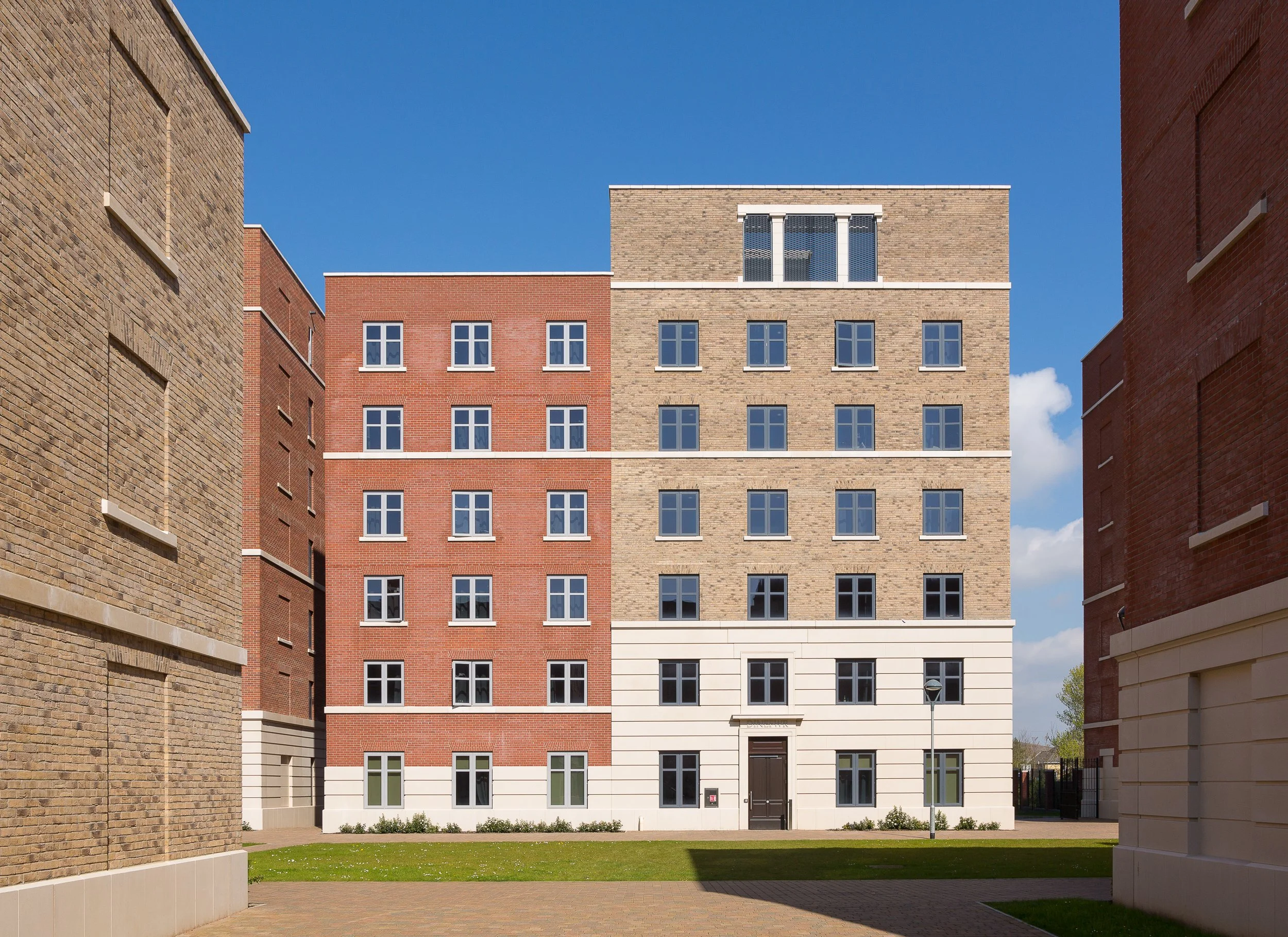 View of a modern multi-story residential building with a mix of red, beige, and white brickwork against a blue sky, framed by other brick buildings on either side.