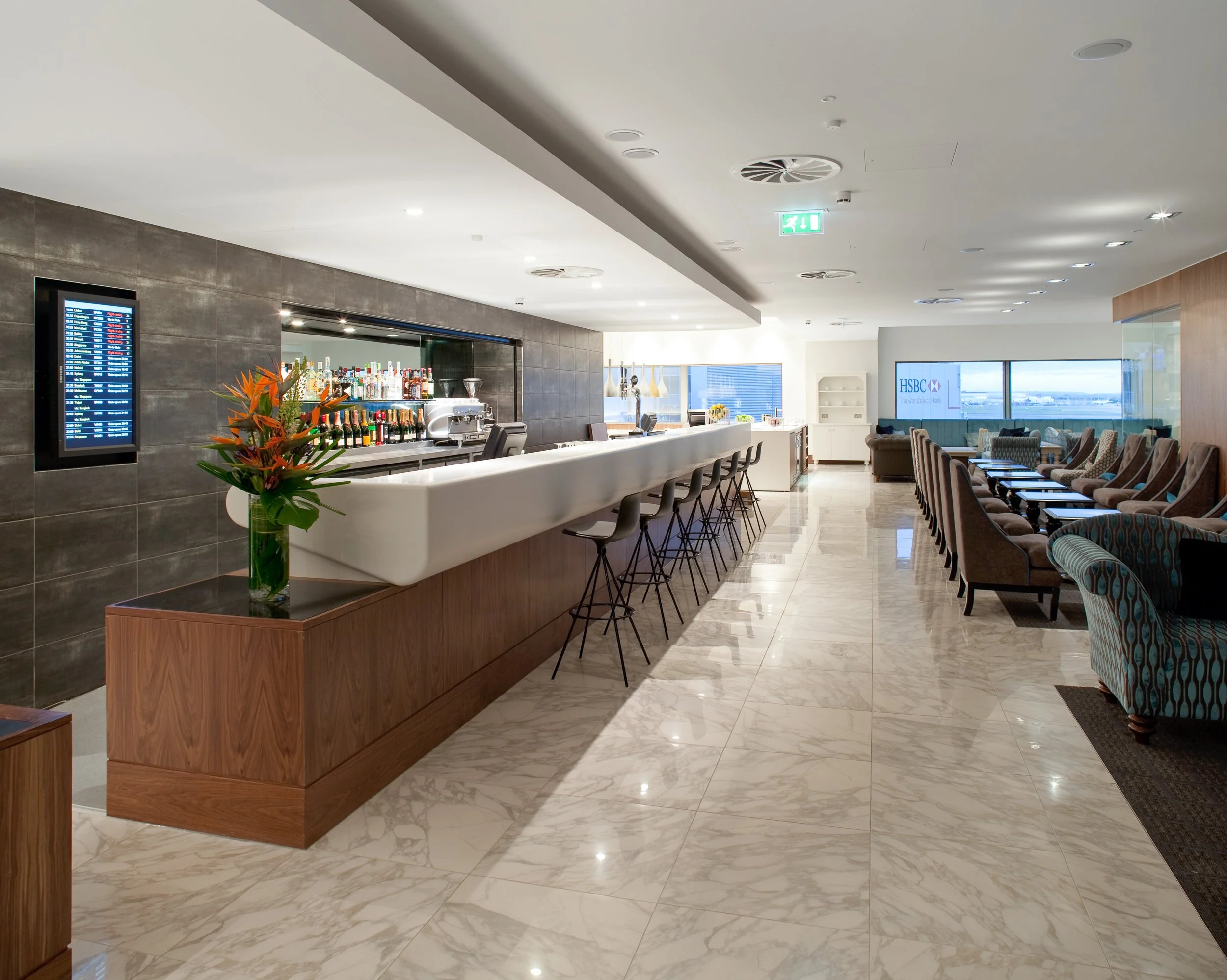 Modern hotel or airport lounge with a bar area, barstools, and a seating lounge with chairs and tables, large windows with a city view.