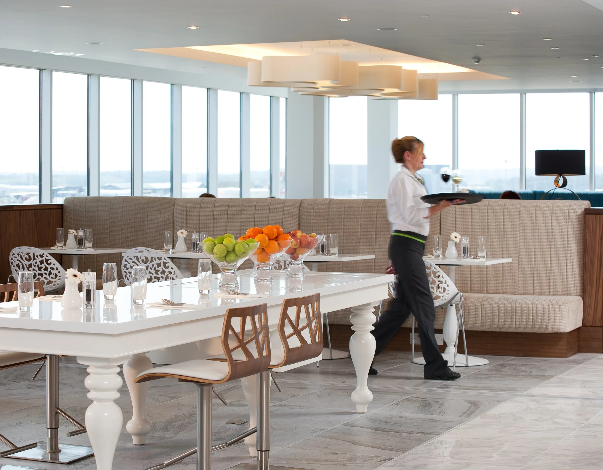 Hotel restaurant with a waitress carrying a tray of beverages near a table with fruit centerpieces, large windows in the background, and modern decor.