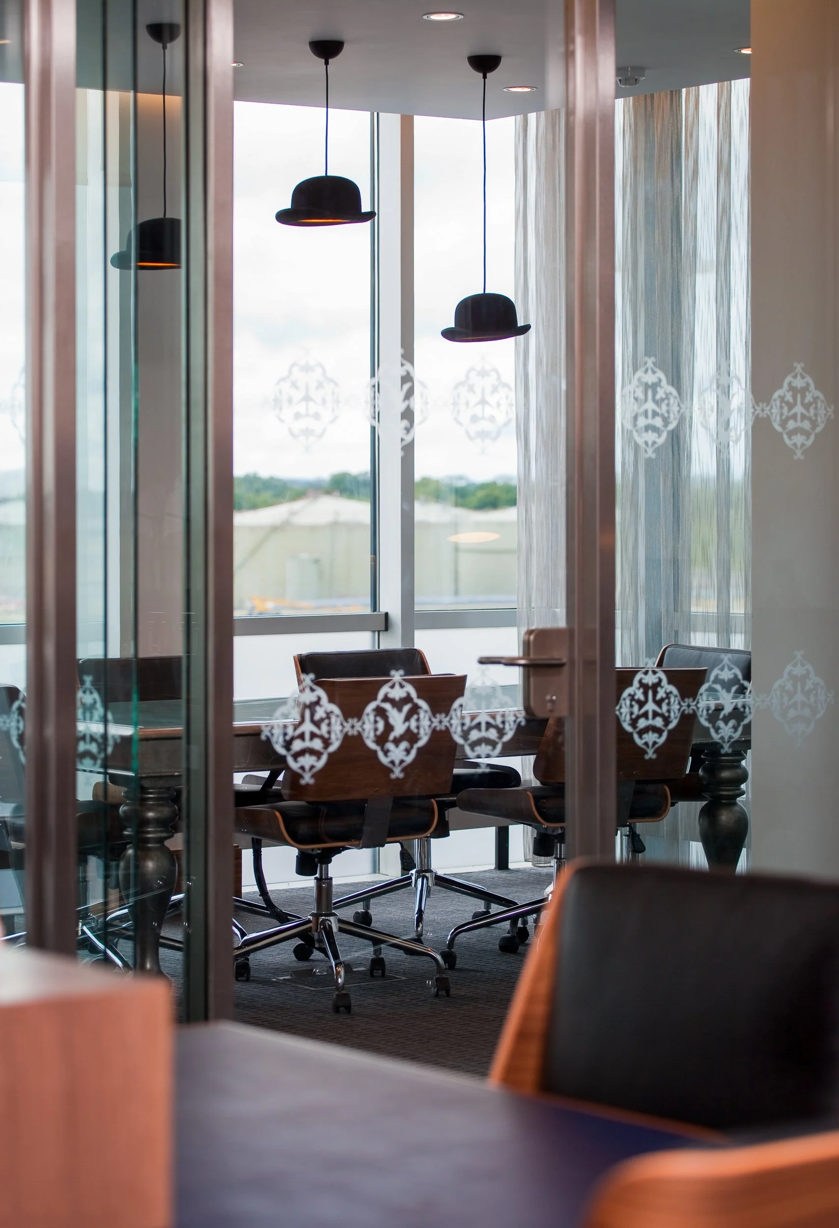 An office conference room seen through glass door with decorative white patterns, featuring a long wooden table, black leather chairs, large windows with a view of the outdoors, and black pendant lights hanging from the ceiling.