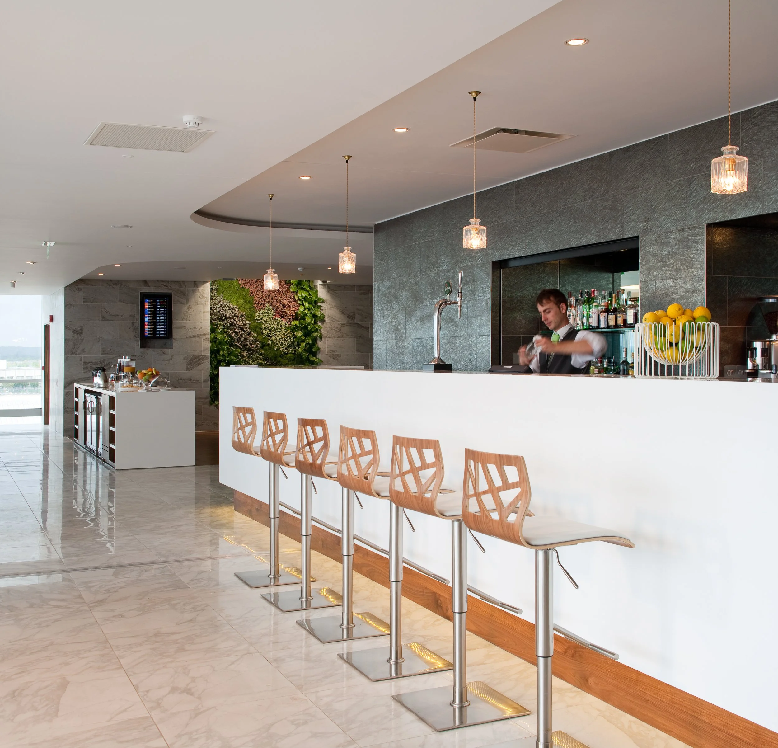 Hotel bar with a bartender, wooden bar stools, modern design, greenery on the wall, and beverage station in the background.