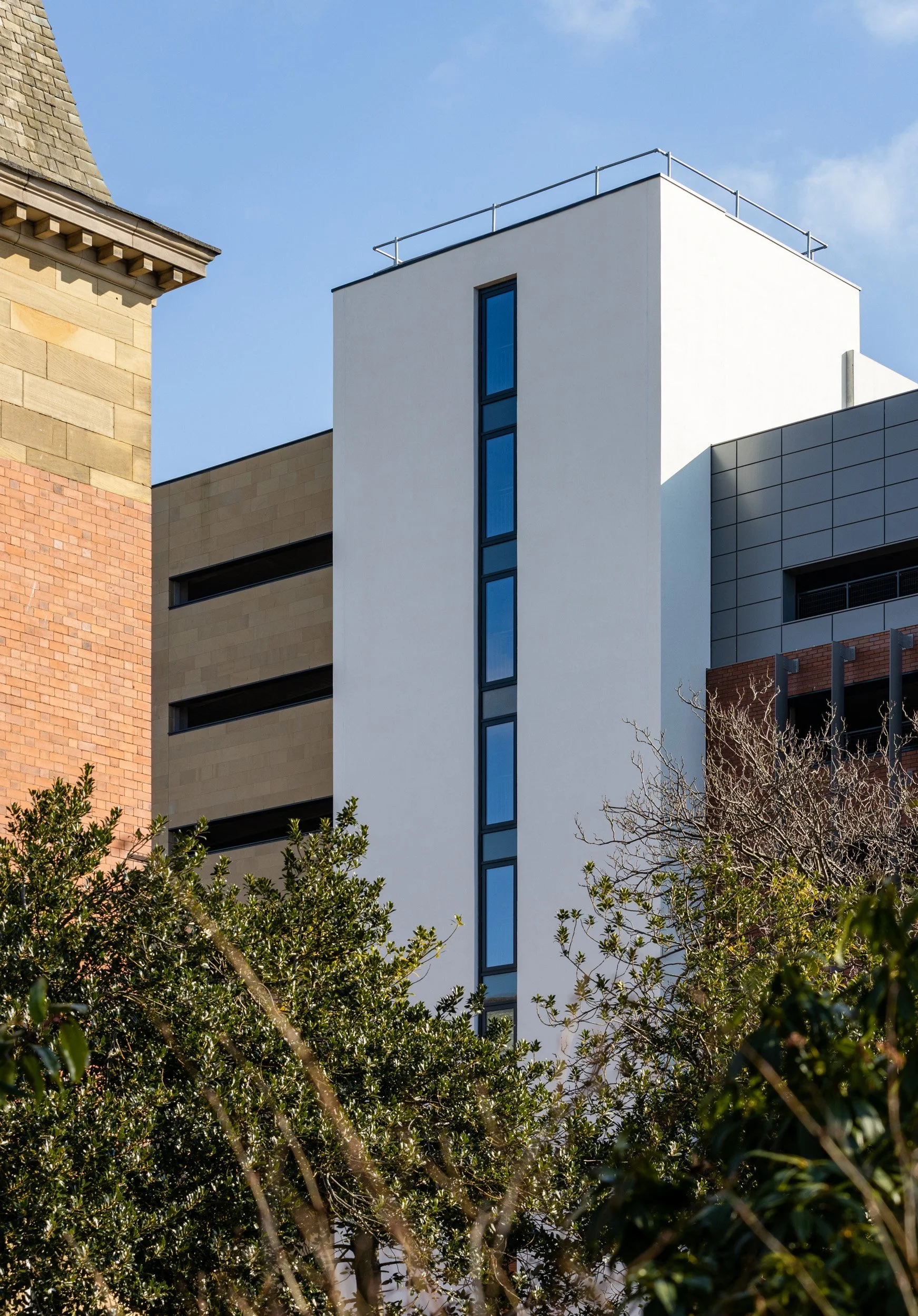 Modern building with white, gray, and beige facades and tall vertical narrow windows, partially obscured by trees in the foreground, under a blue sky.