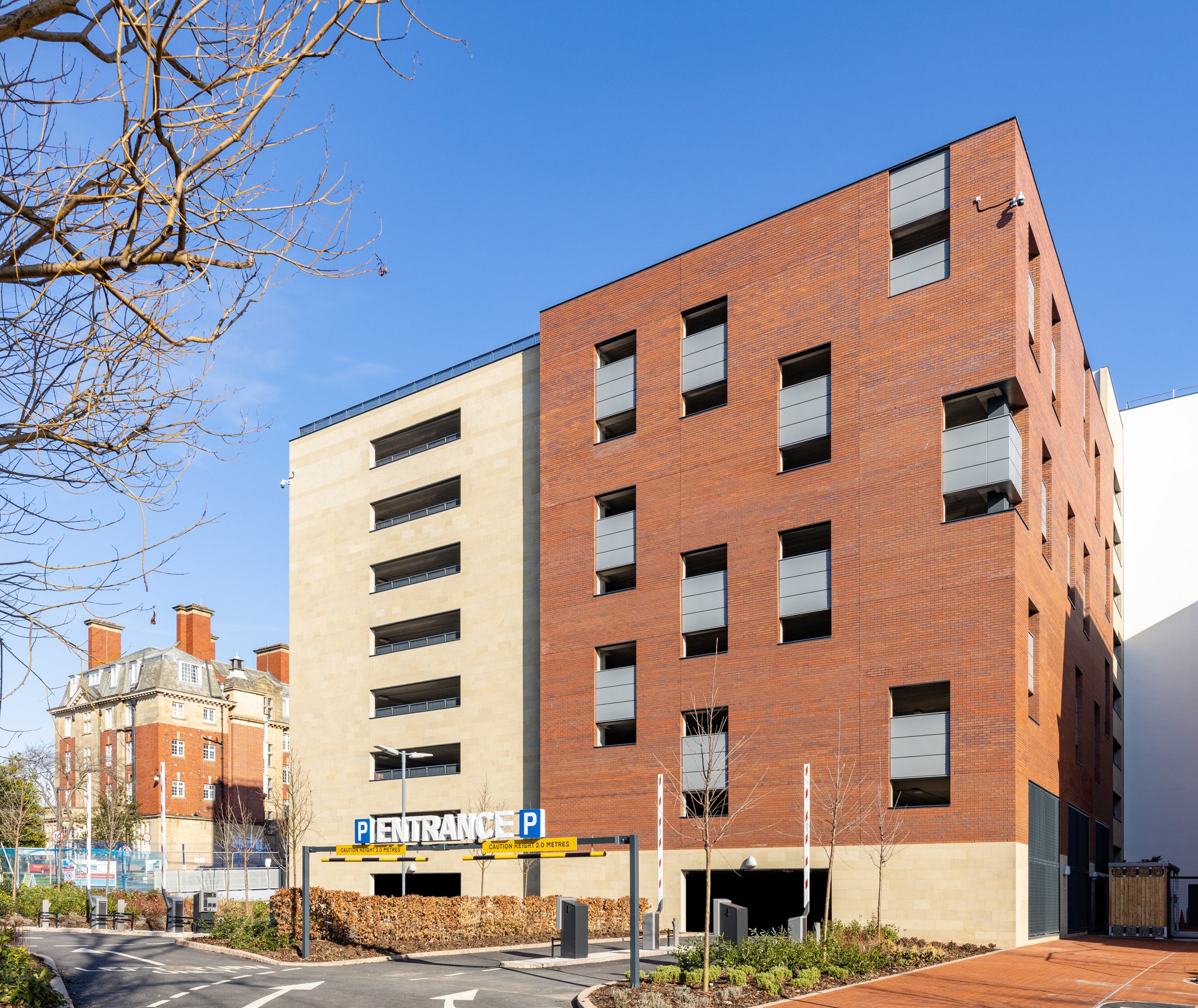 Modern multi-story residential or commercial building with a mix of red brick and light-colored facade, multiple windows and balconies, and an underground parking entrance with parking signs and a small landscaped area in the foreground.