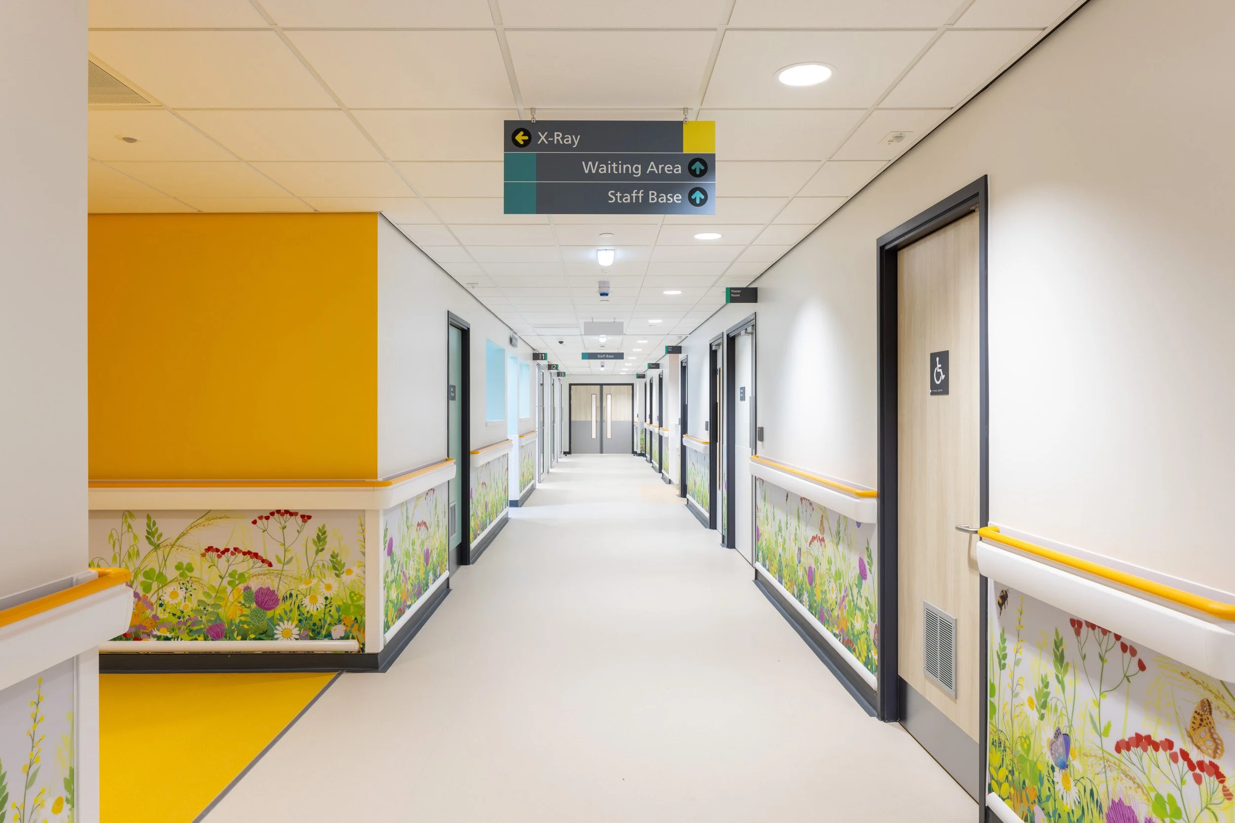Hospital corridor with doors, colorful floral decorations, and overhead signs for X-Ray, Waiting Area, and Staff Base.