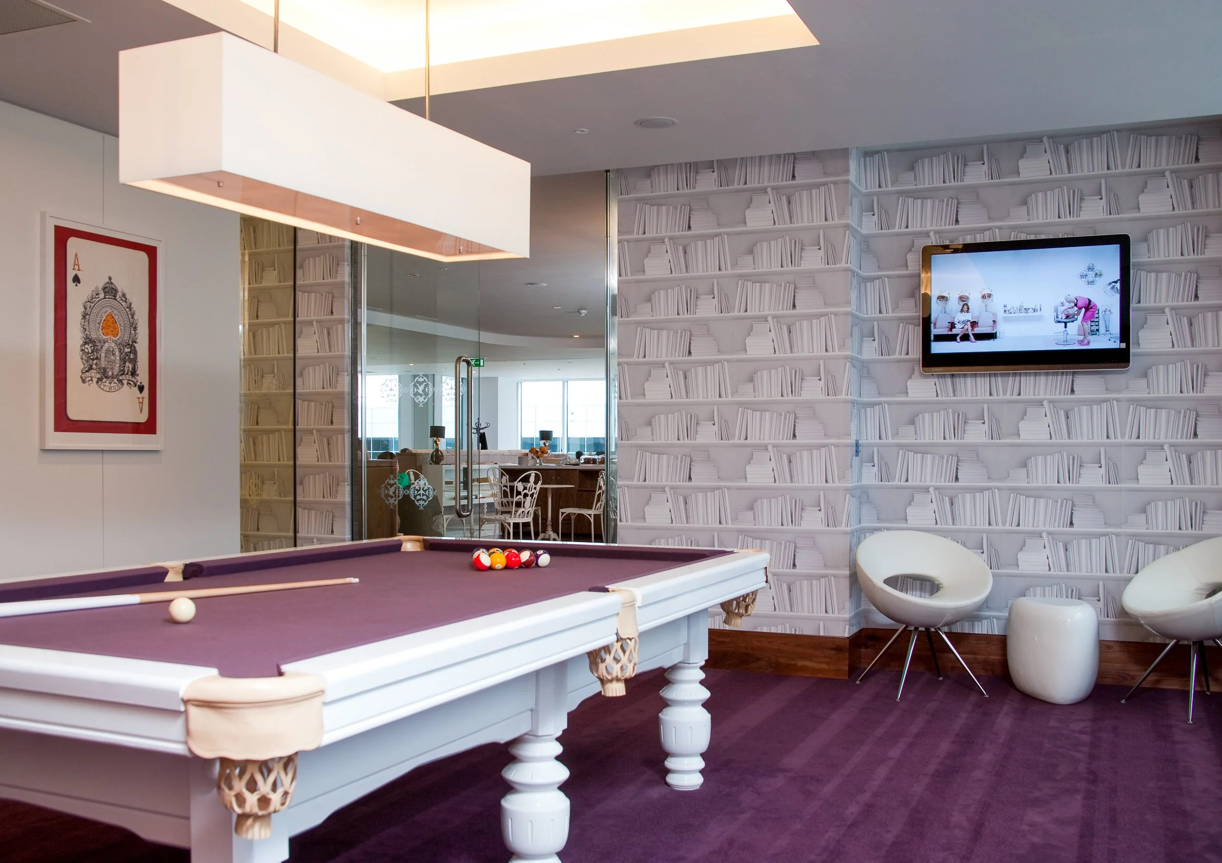 A room with a pool table with purple felt, a large white rectangular light fixture, a wall with a bookshelf wallpaper pattern, a flat screen TV displaying people in a living room, and modern seating including white chairs and a small white ottoman.