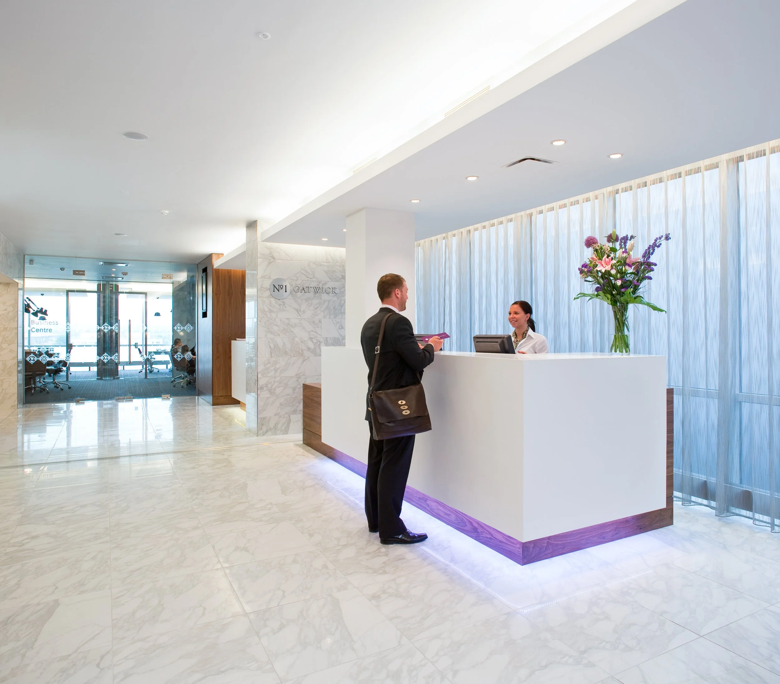 A man in a suit checking in at a hotel reception desk with a smiling female clerk, a large flower arrangement on the desk, and a modern, bright lobby with sheer curtains and marble floors.