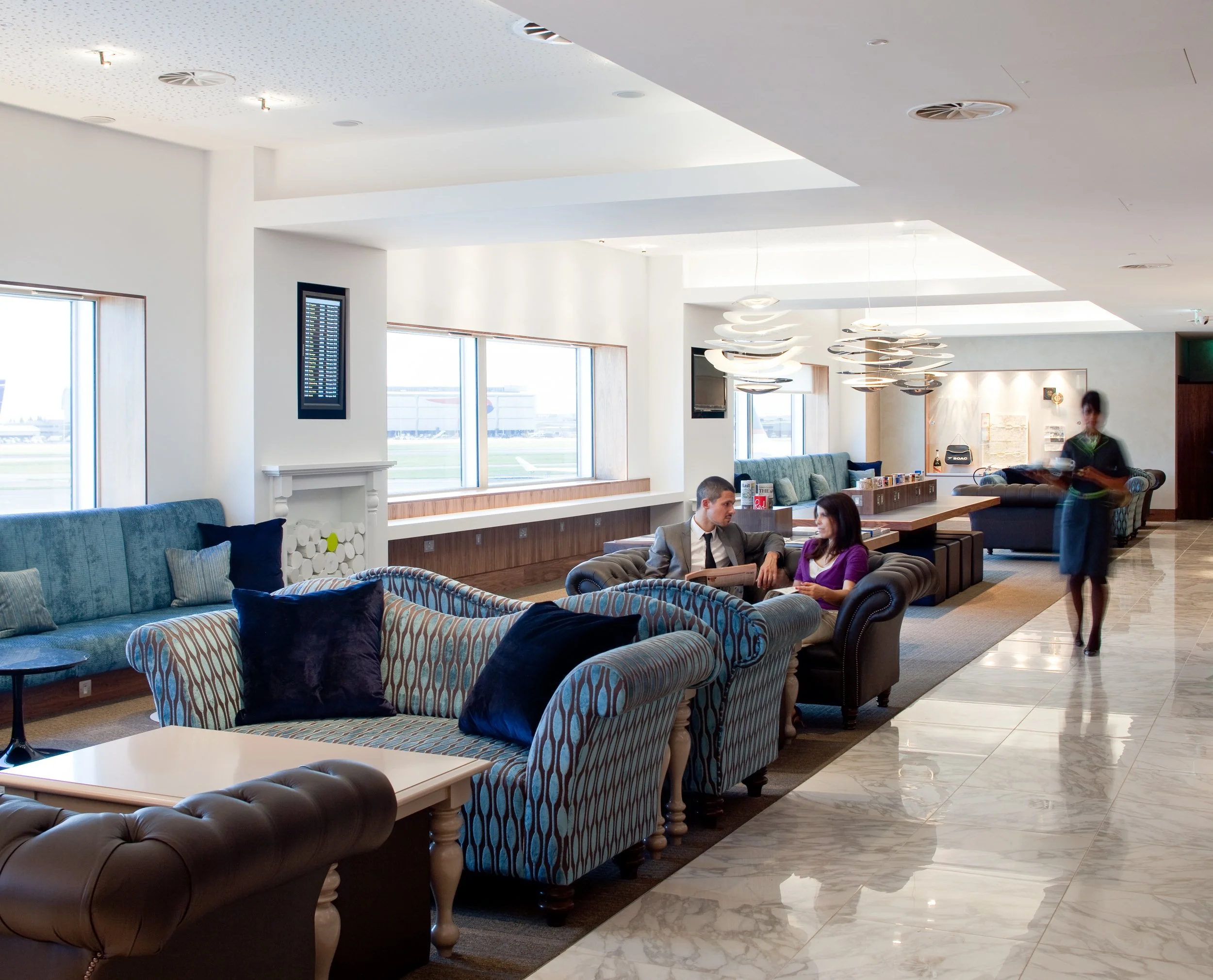 A spacious airport lounge with large windows, decorated with modern furniture including striped and solid colored sofas and chairs. Two people are seated and talking, while a staff member approaches holding a tray. The lounge features decorative ligh