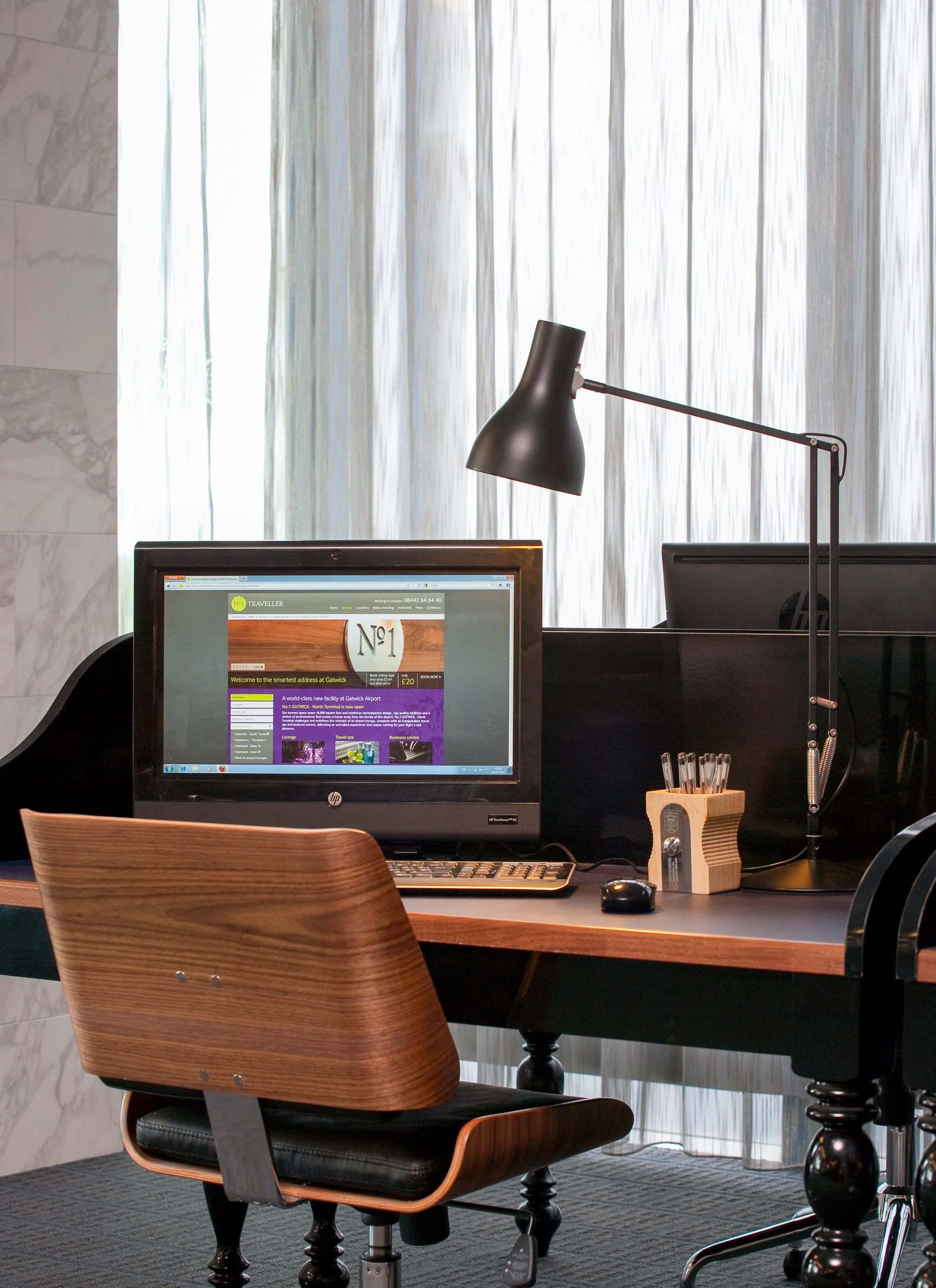 An office workspace with a computer, keyboard, mouse, desk lamp, and wooden pen holder with pens, positioned in front of a window with sheer curtains.