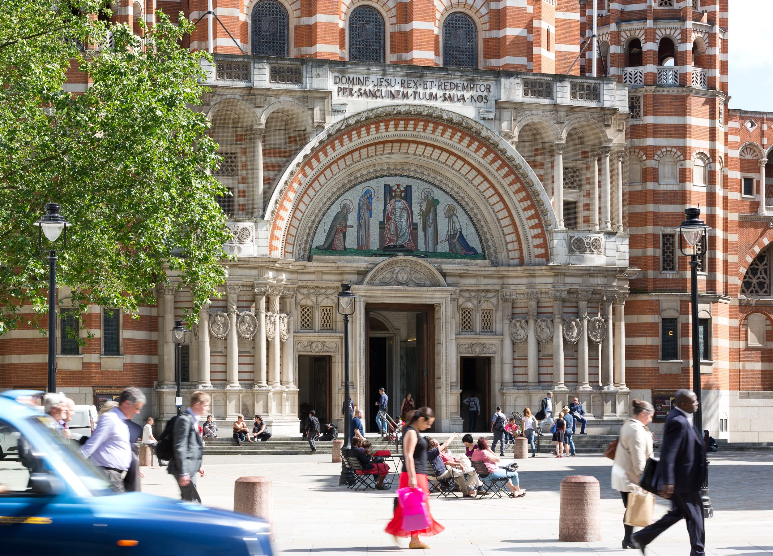 People walking and sitting in front of a historic church with ornate architecture, red and white brick, and mosaic artwork above the entrance.