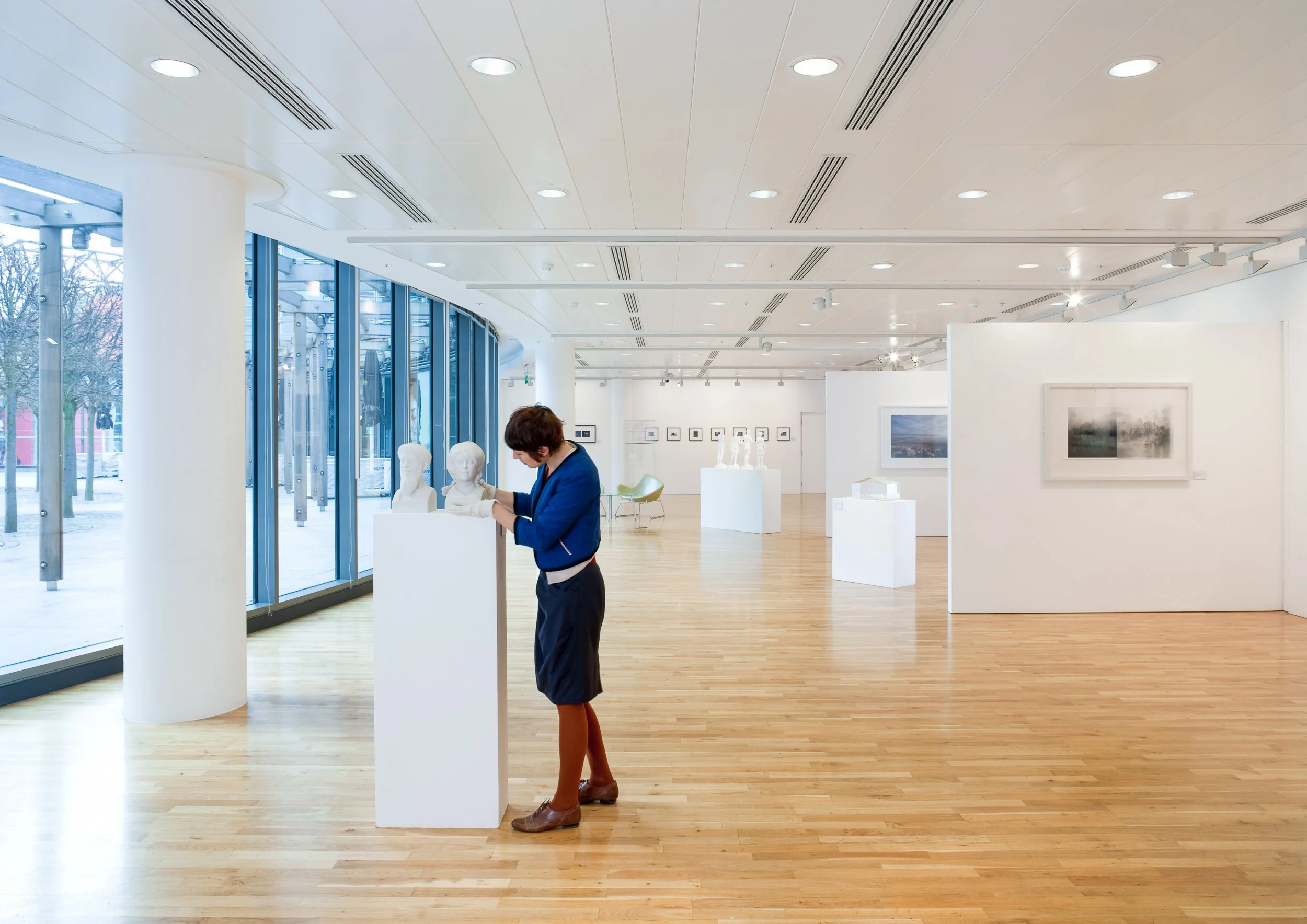 An art gallery with wooden floors, white walls, and large glass windows. A woman in a blue jacket and brown boots is inspecting bust sculptures on a white pedestal. The gallery contains various artworks, including paintings and sculptures, displayed 