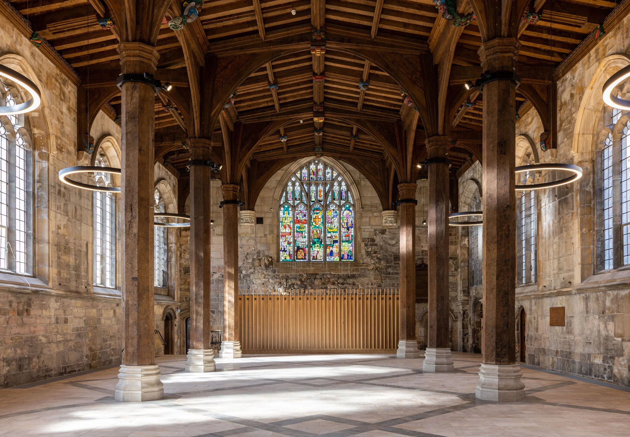 Interior of a historic church or cathedral with stone walls, large stained glass window at the end, wooden ceiling with exposed beams, circular modern pendant lights, and tall arched windows along the sides.