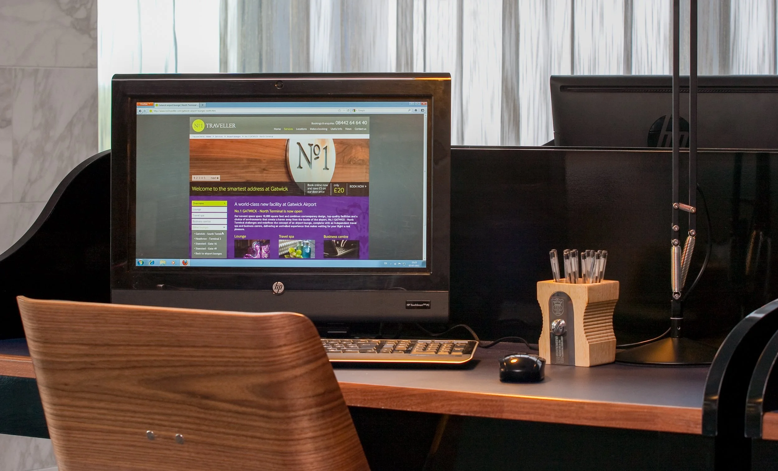 An office desk with a computer monitor displaying a webpage about Gatwick Airport. The desk has a wooden chair in front, a keyboard, a mouse, and a wooden pen holder with pens. There is also a lamp on the right side of the desk and a black partition 