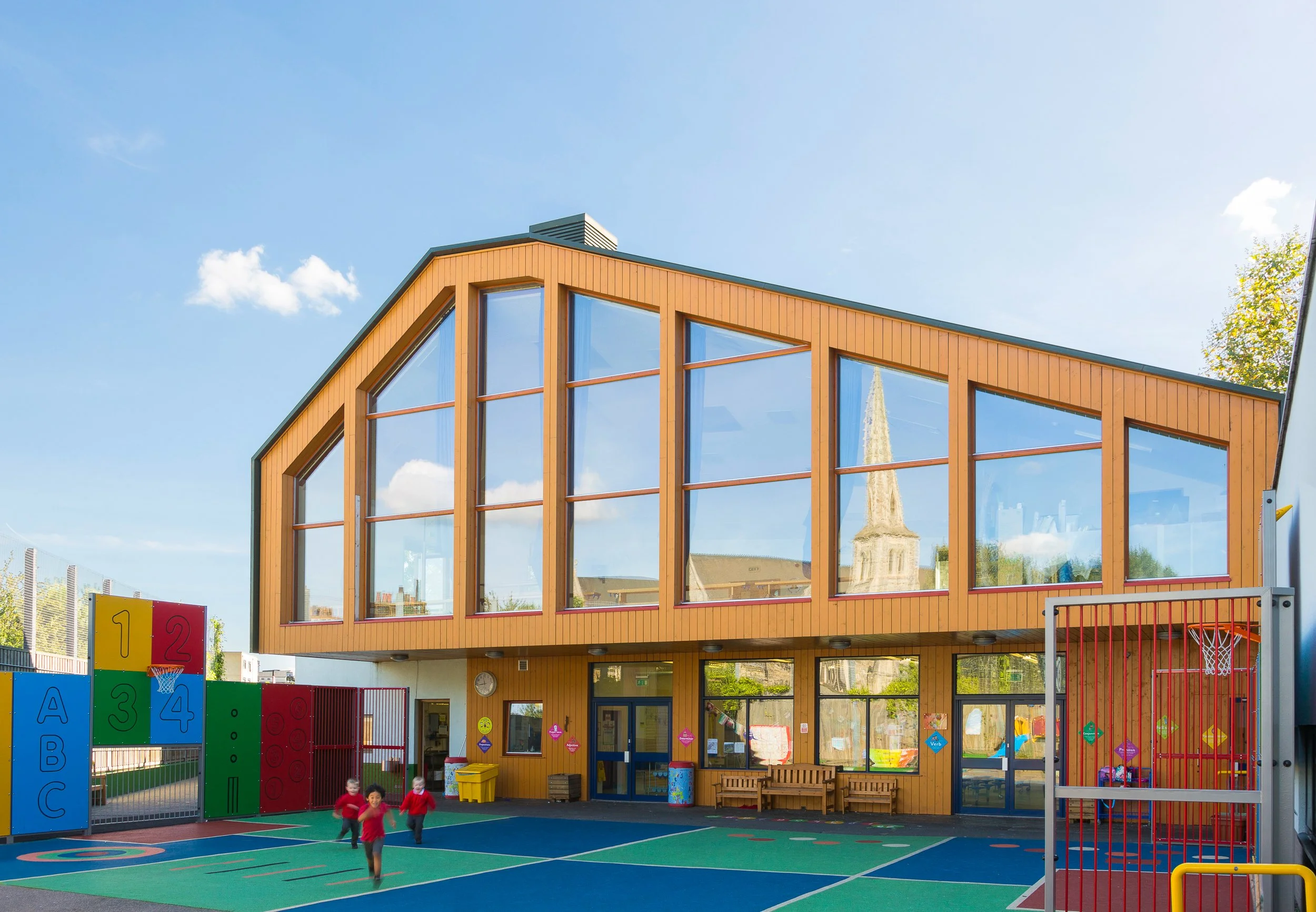 A modern two-story school building with large glass windows and wooden panels, featuring a colorful outdoor playground with a basketball hoop and children playing beneath a blue sky.