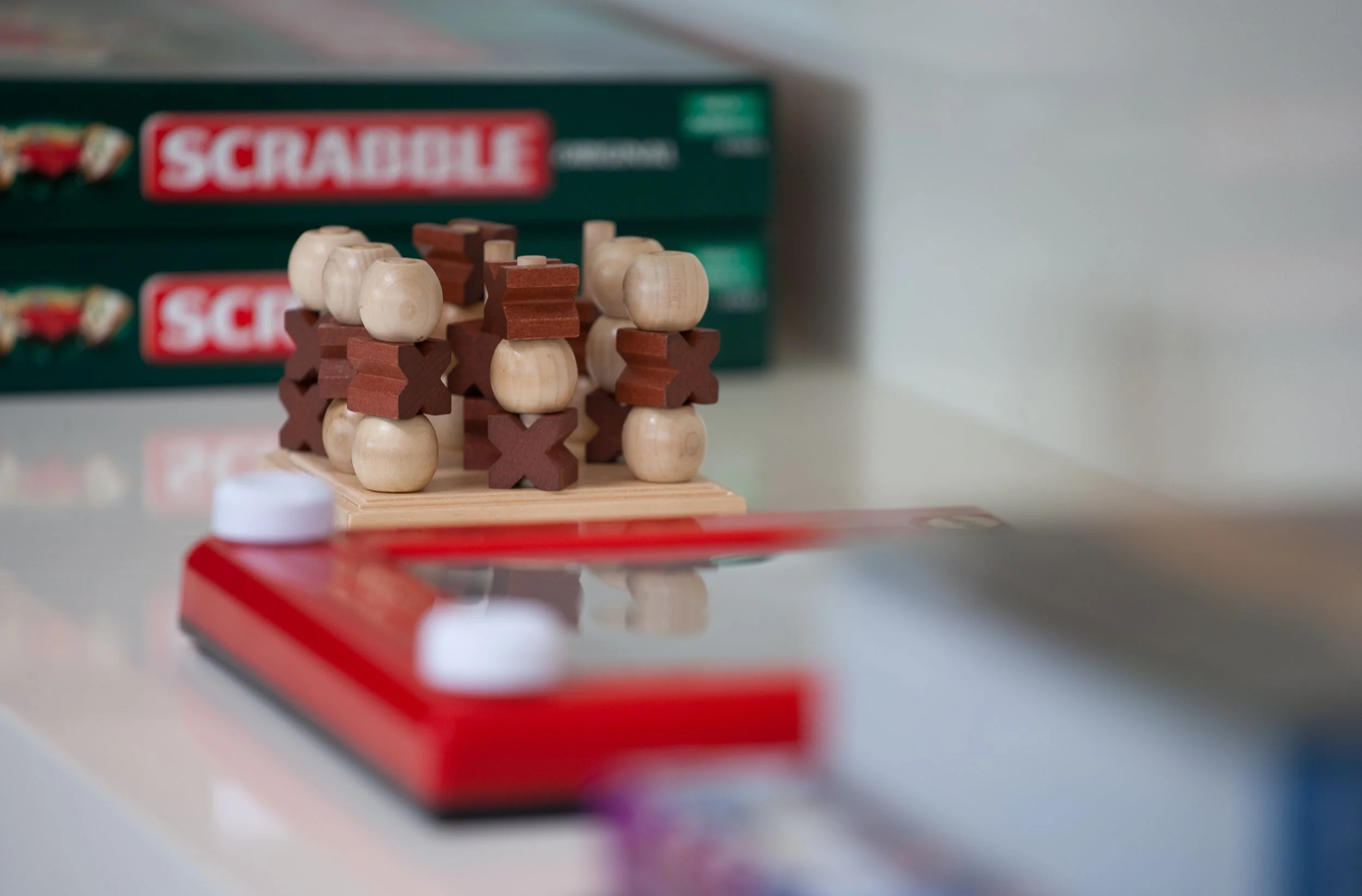 Close-up of a wooden tic-tac-toe game with some X and O pieces, with Scrabble game boxes in the background on a white table.