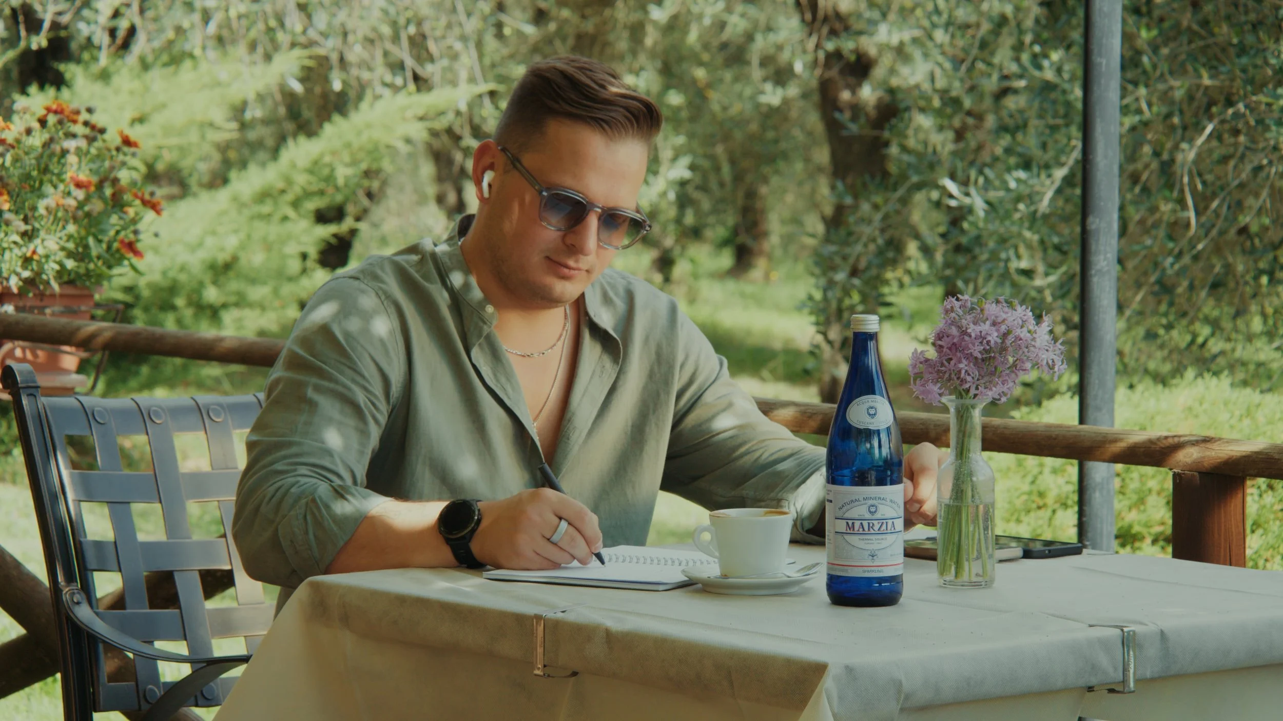Man sat at a table outside, writing in a journal with a bottle of Marzia water in front of him